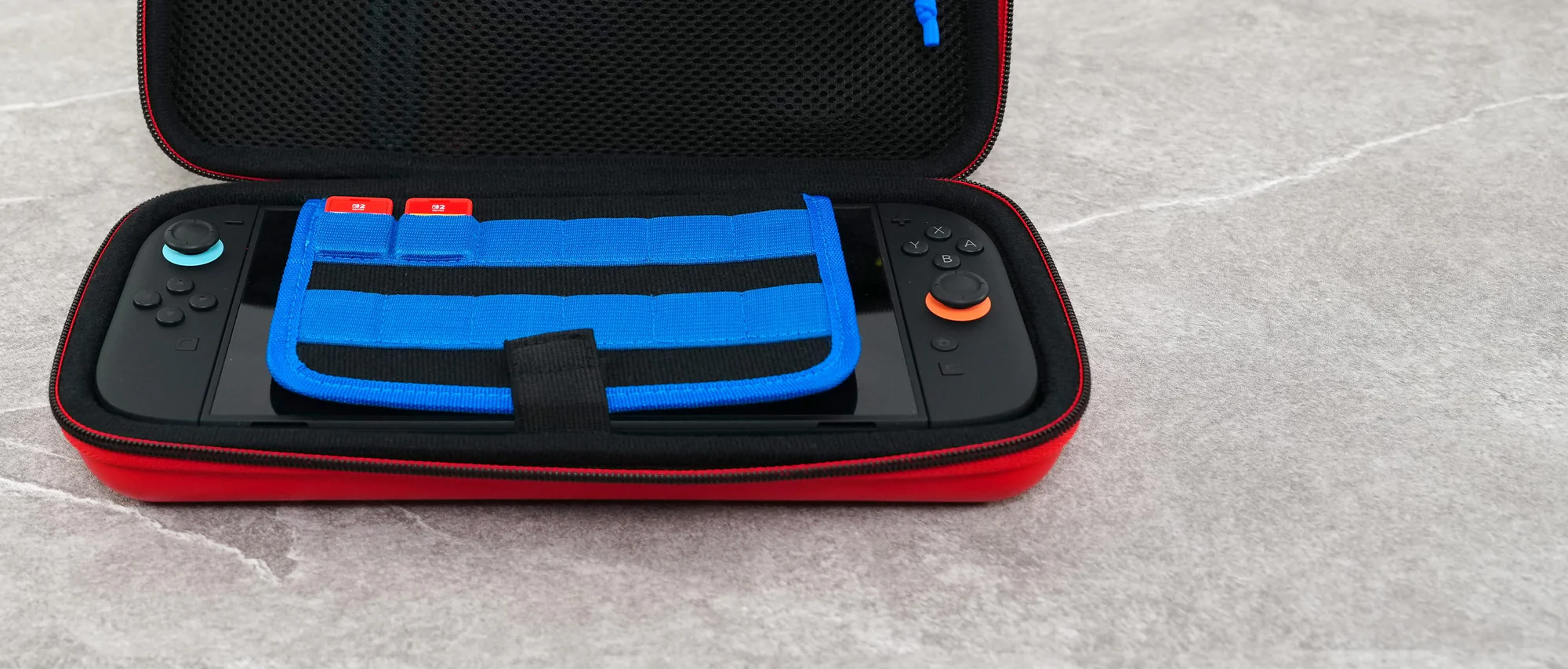 Un Nintendo Switch nero in una custodia rossa con un accessorio blu. La custodia è aperta su una superficie grigia.