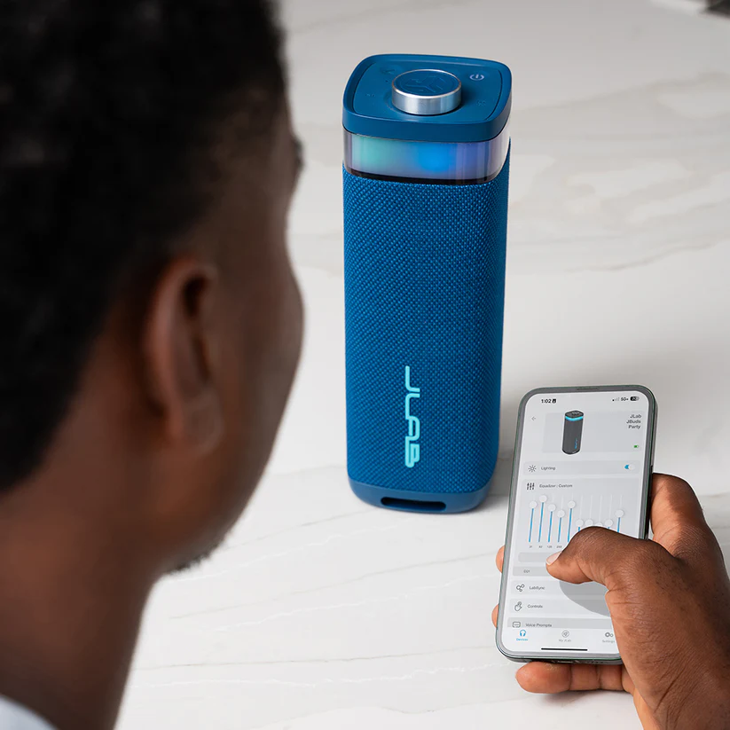Eine Person steuert einen blauen JLab-Lautsprecher mit einem Smartphone. Der Lautsprecher befindet sich neben der Person. Weiße Oberfläche.