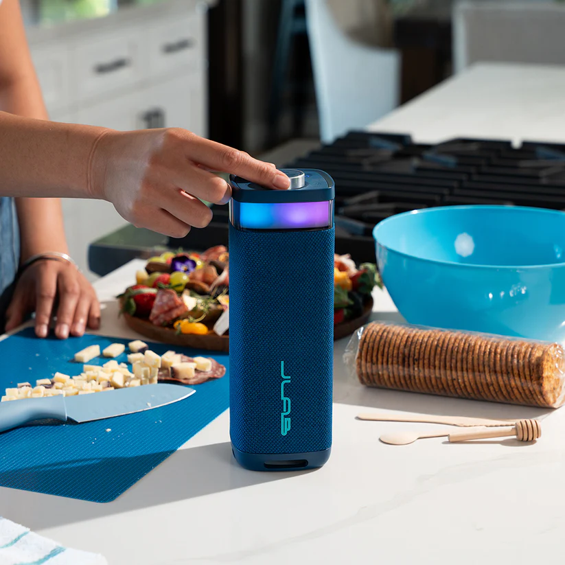 Eine Person drückt auf einen Knopf an einem blauen Lautsprecher. Ein Schneidebrett mit Käse und Essen, eine Schüssel und Cracker sind in der Nähe.