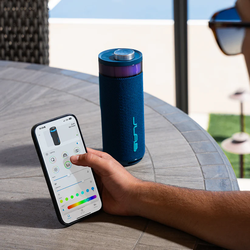 Eine Person steuert einen blauen Lautsprecher mit einem Telefon. Das Telefon zeigt eine Steuerungs-App. Der Lautsprecher steht auf einem Steintisch.