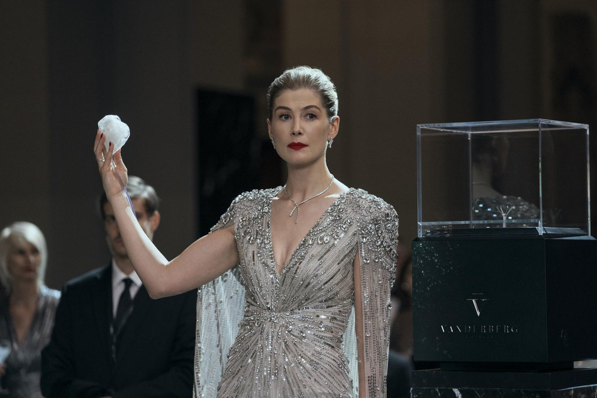 Eine Frau in einem eleganten Kleid hält einen Diamanten. Ein Diamant befindet sich in einer Glasvitrine. Menschen sind im Hintergrund.