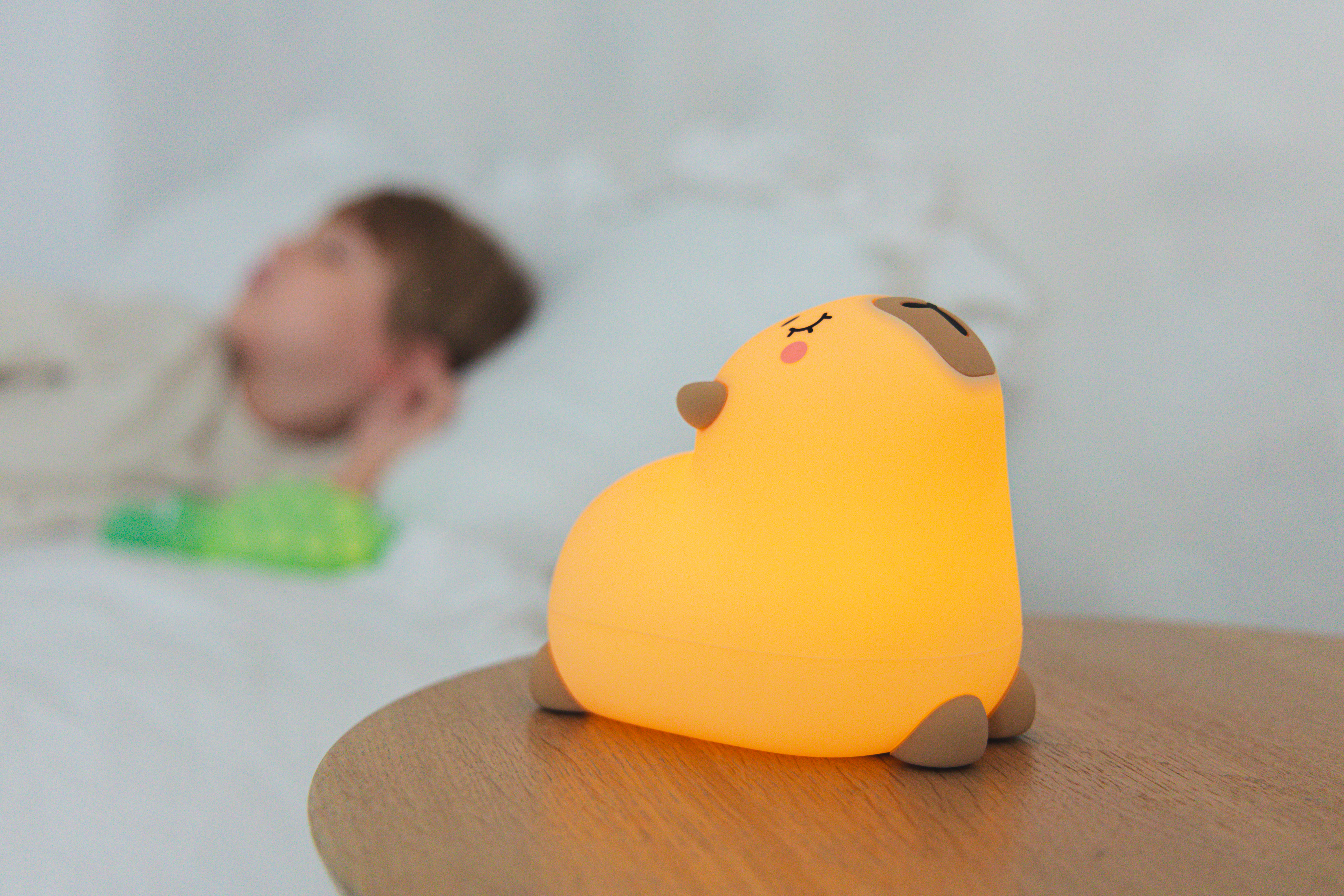 Świecąca na żółto lampa w kształcie psa na drewnianym stole. Osoba leży w łóżku w rozmytym tle.