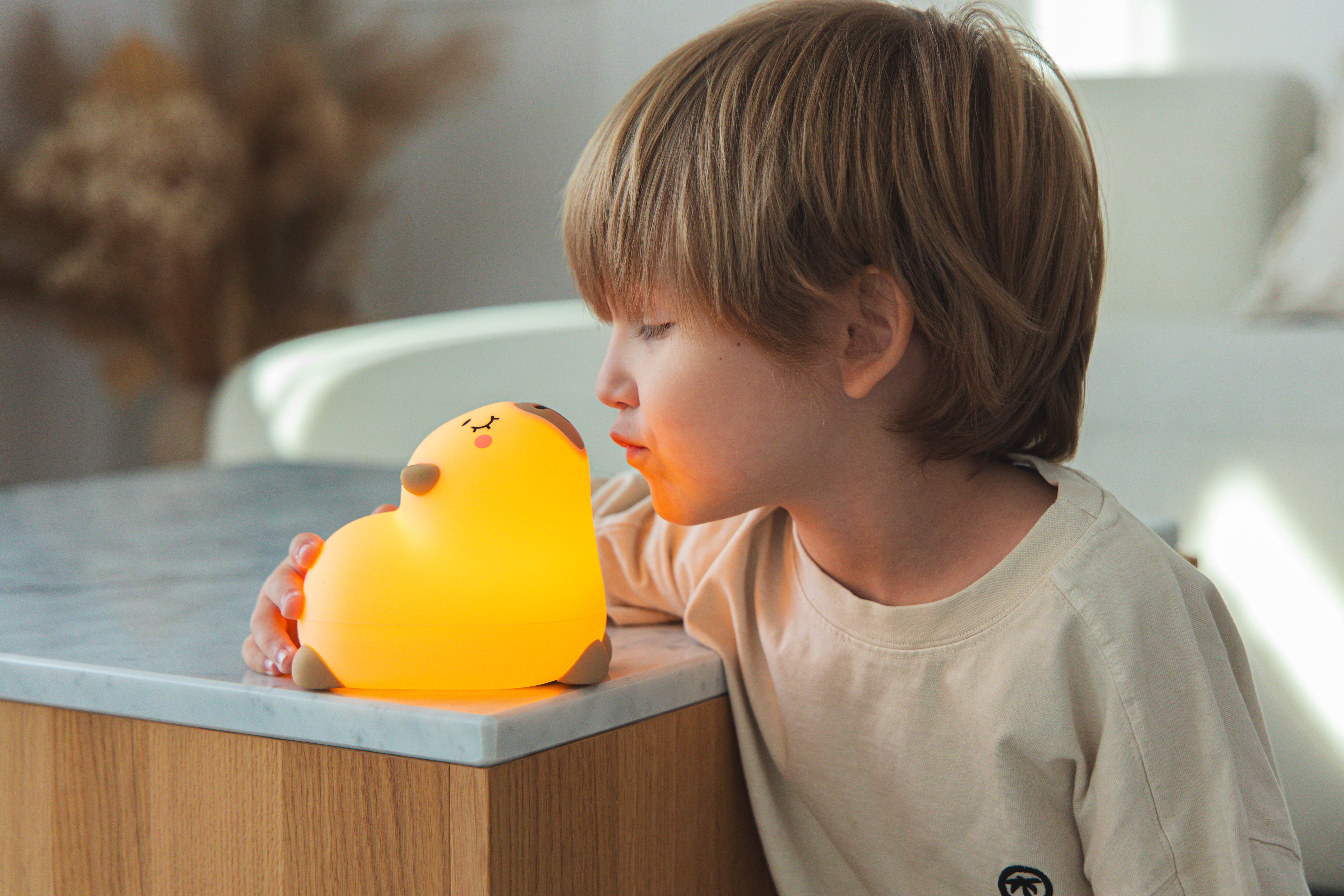 Dziecko dmucha na żółtą lampę w kształcie serca. Lampa stoi na marmurowej powierzchni. Dziecko ma na sobie jasną koszulę.