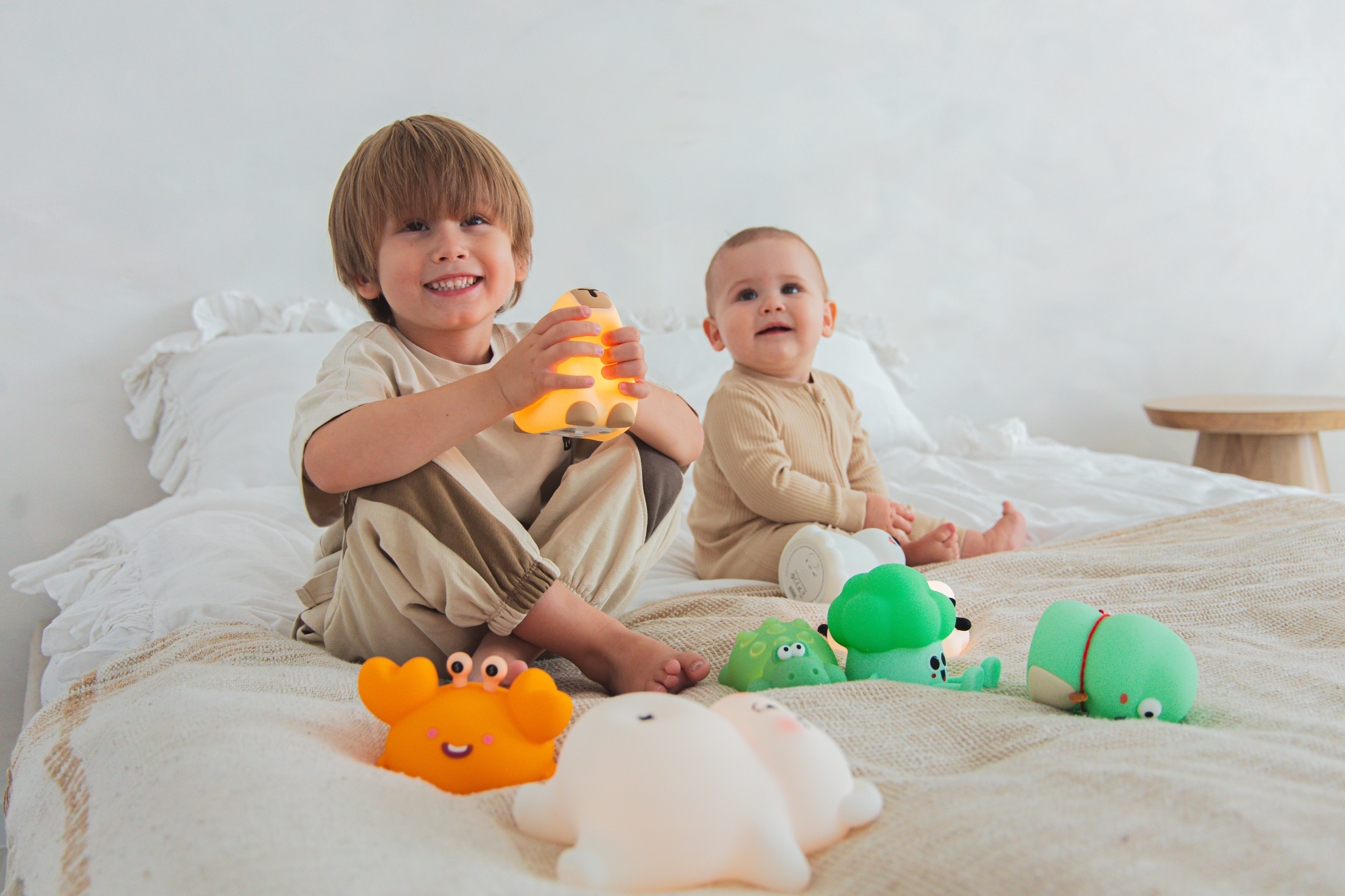 Dwójka dzieci na łóżku z zabawkami. Jedno trzyma świecącą lampę w kształcie misia. Inne zabawki mają kształt zwierząt.