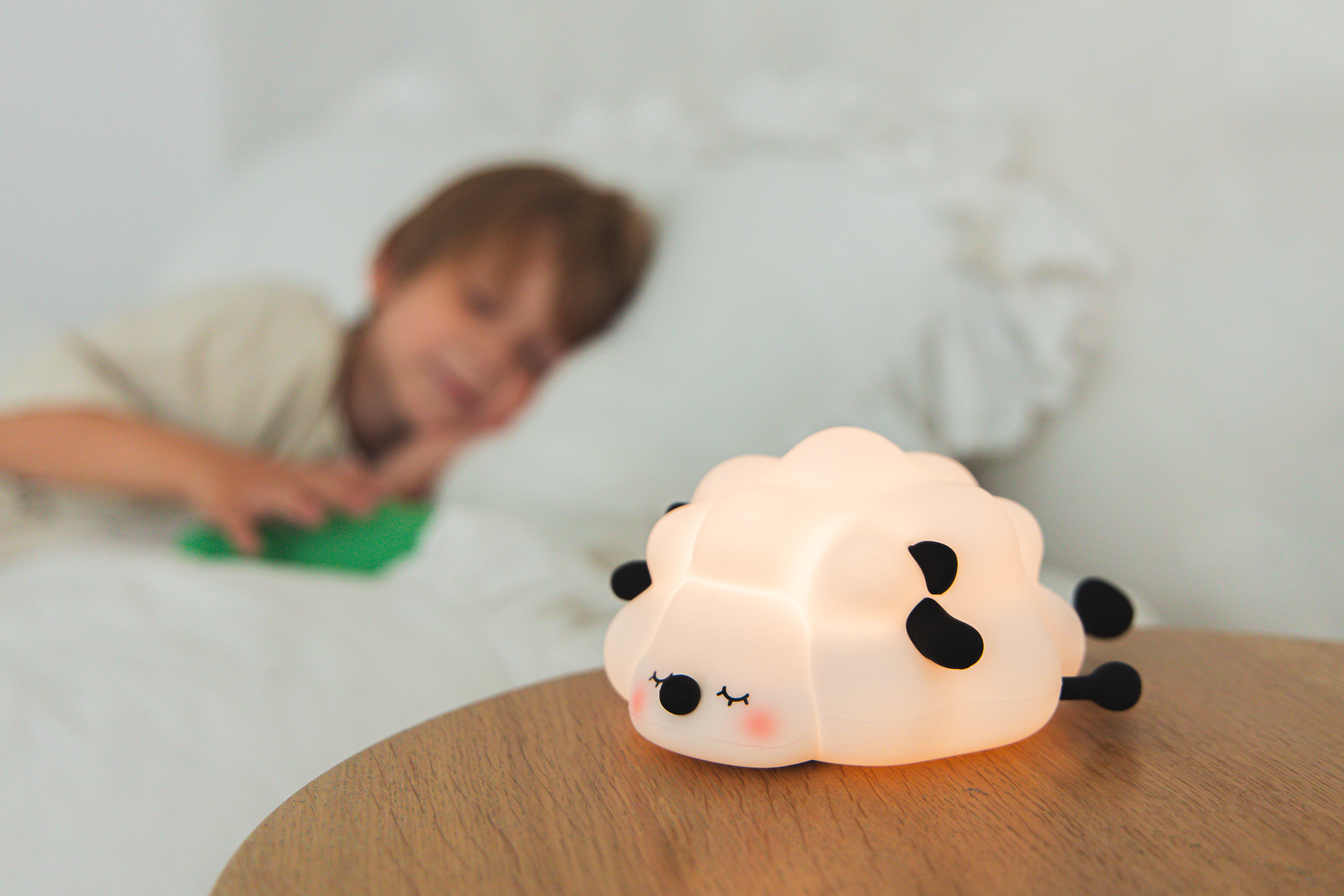 Świecąca lampka nocna w kształcie owcy stoi na drewnianym stole. Dziecko odpoczywa w rozmytym tle.