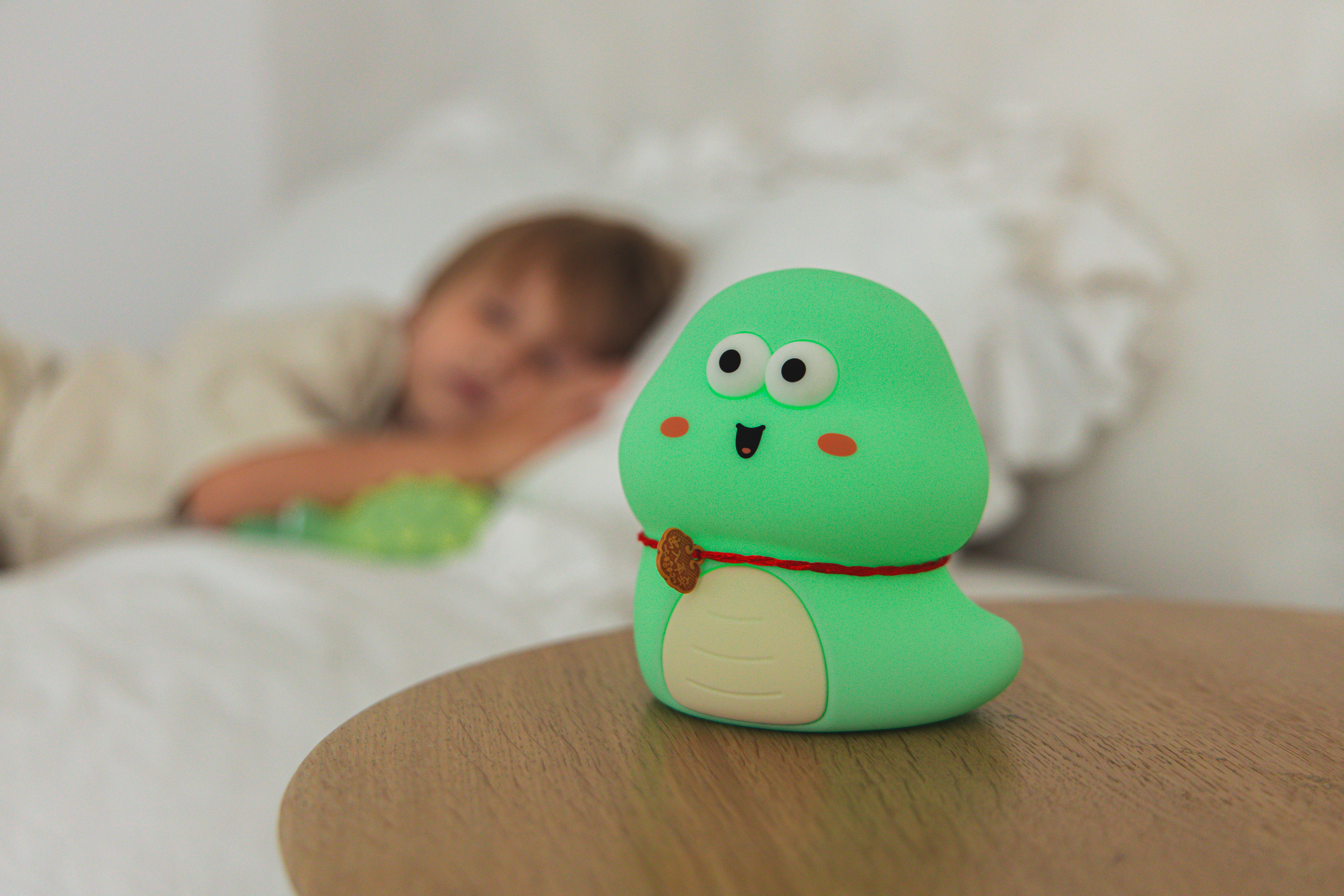 Zielony dinozaur zabawka na drewnianym stole. W tle śpi dziecko. Zabawka ma czerwony naszyjnik.