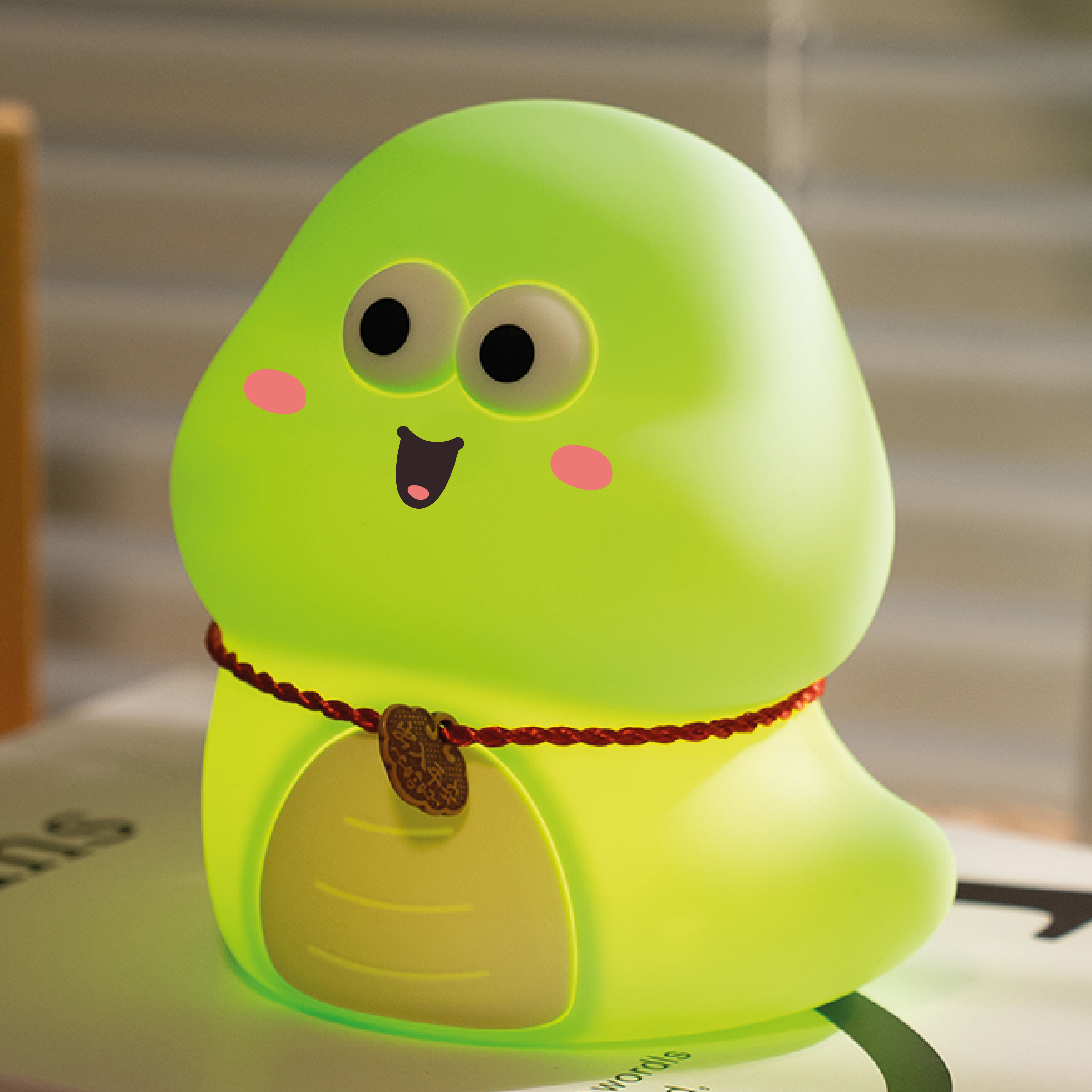 Zielona lampa w kształcie dinozaura z uśmiechniętą twarzą, różowymi policzkami i czerwonym naszyjnikiem. Leży na książce.
