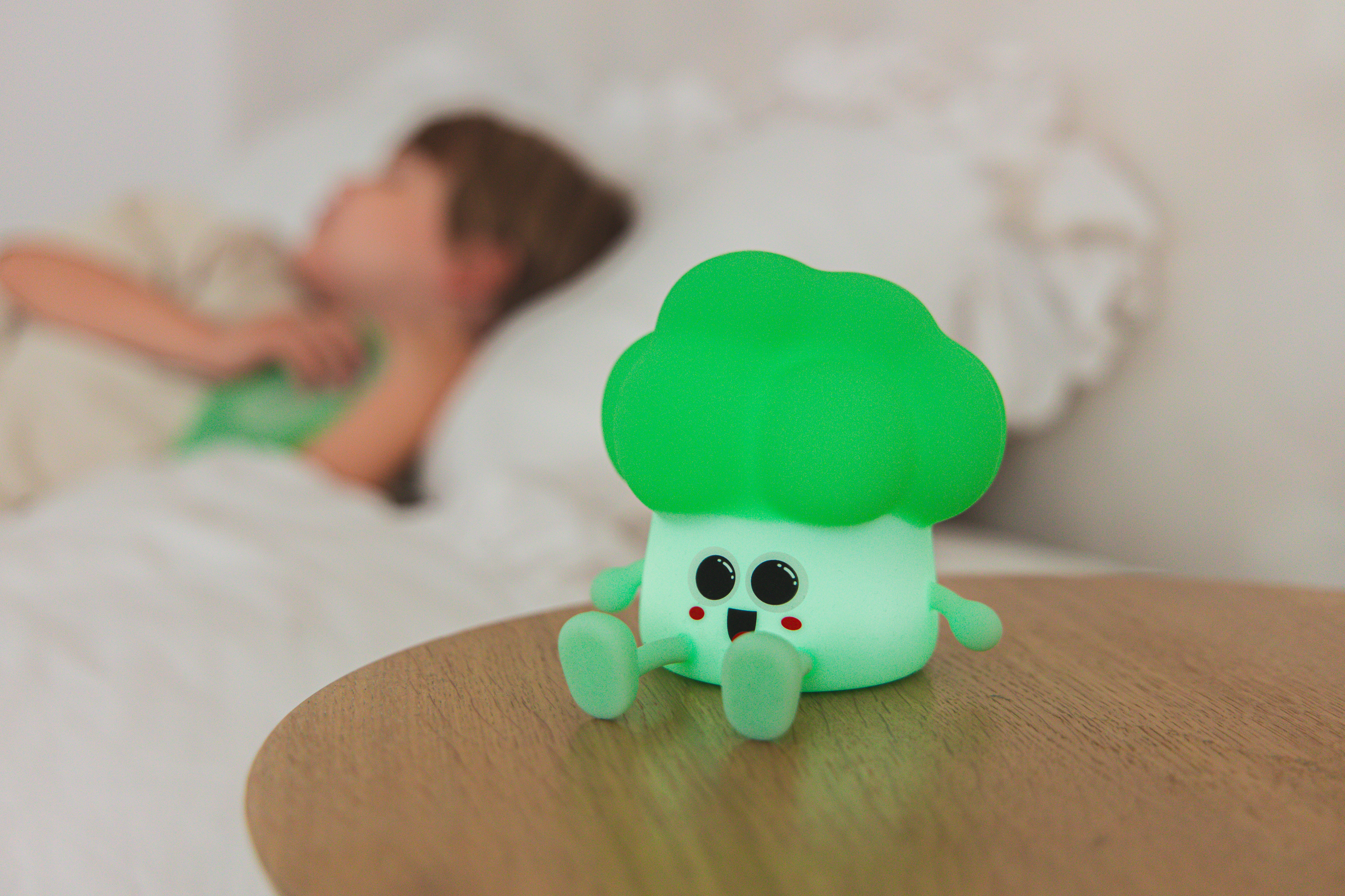 Zielona lampka nocna w kształcie brokułu stoi na stole. Rozmazane dziecko śpi w tle.