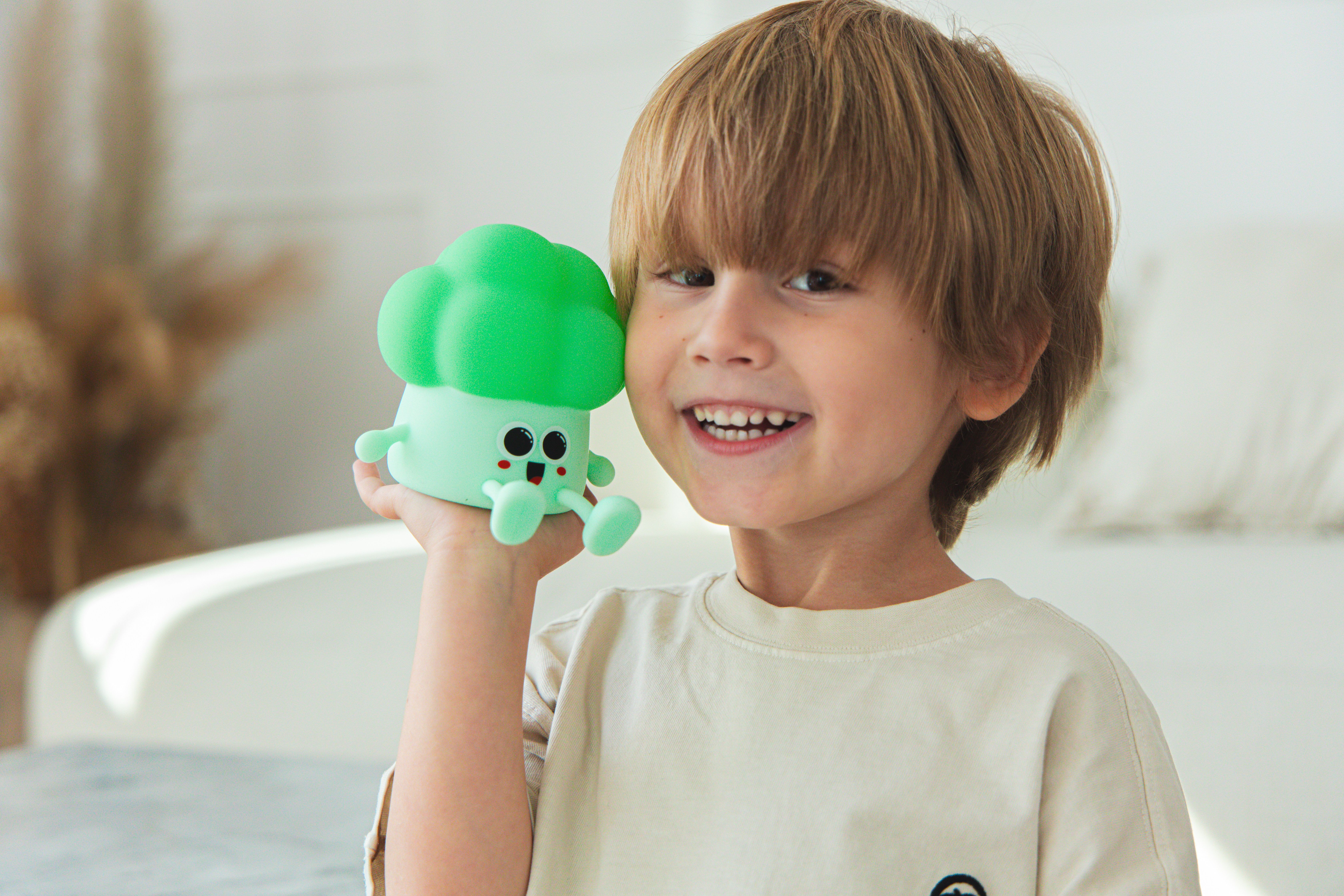 Uśmiechnięty chłopiec trzyma zieloną zabawkę brokuł. Tło jest rozmyte.