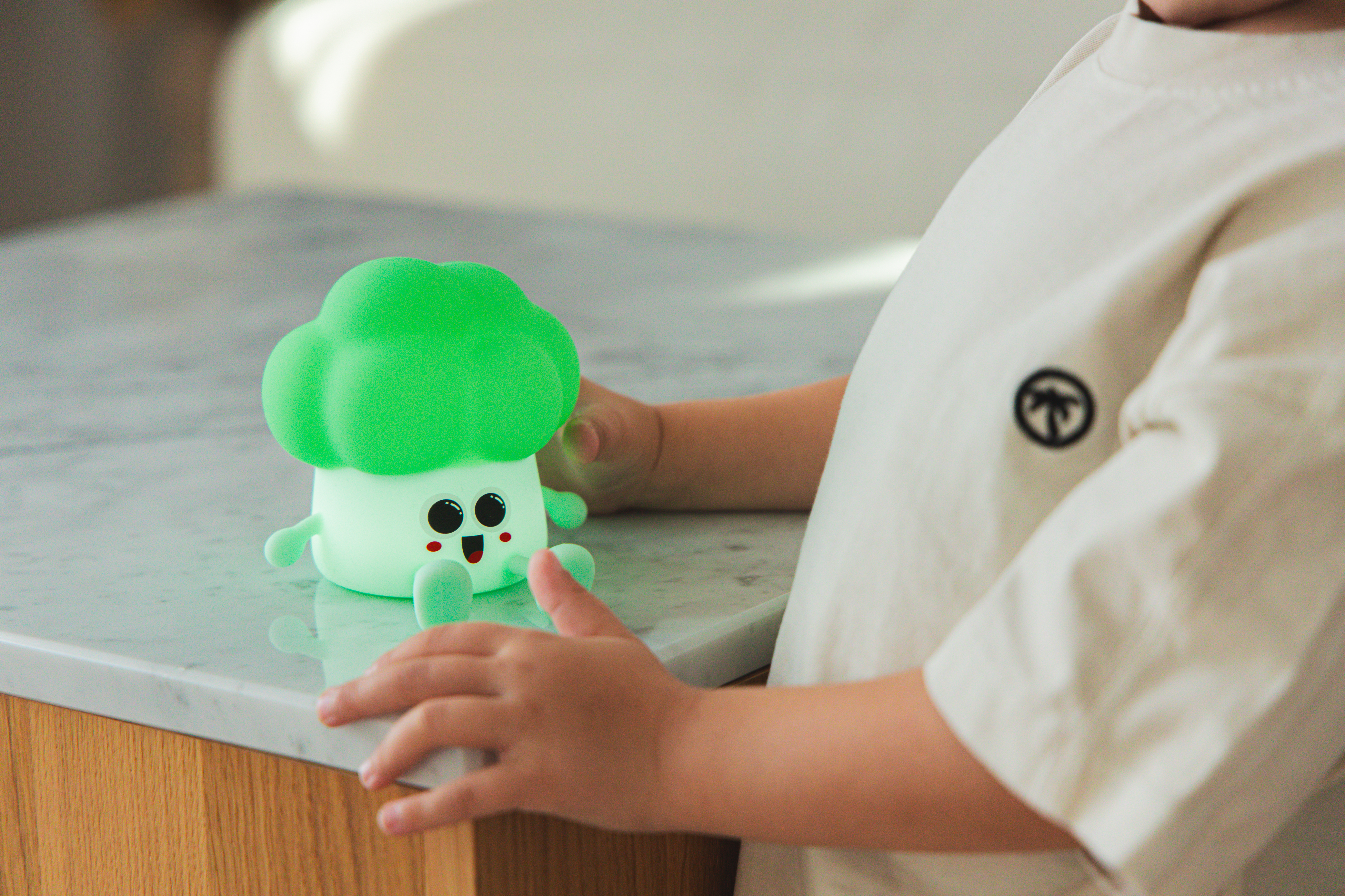 Ręce i koszulka dziecka kadrują uśmiechniętą zabawkę w kształcie brokuła. Zabawka stoi na marmurowej powierzchni. Koszulka ma logo.