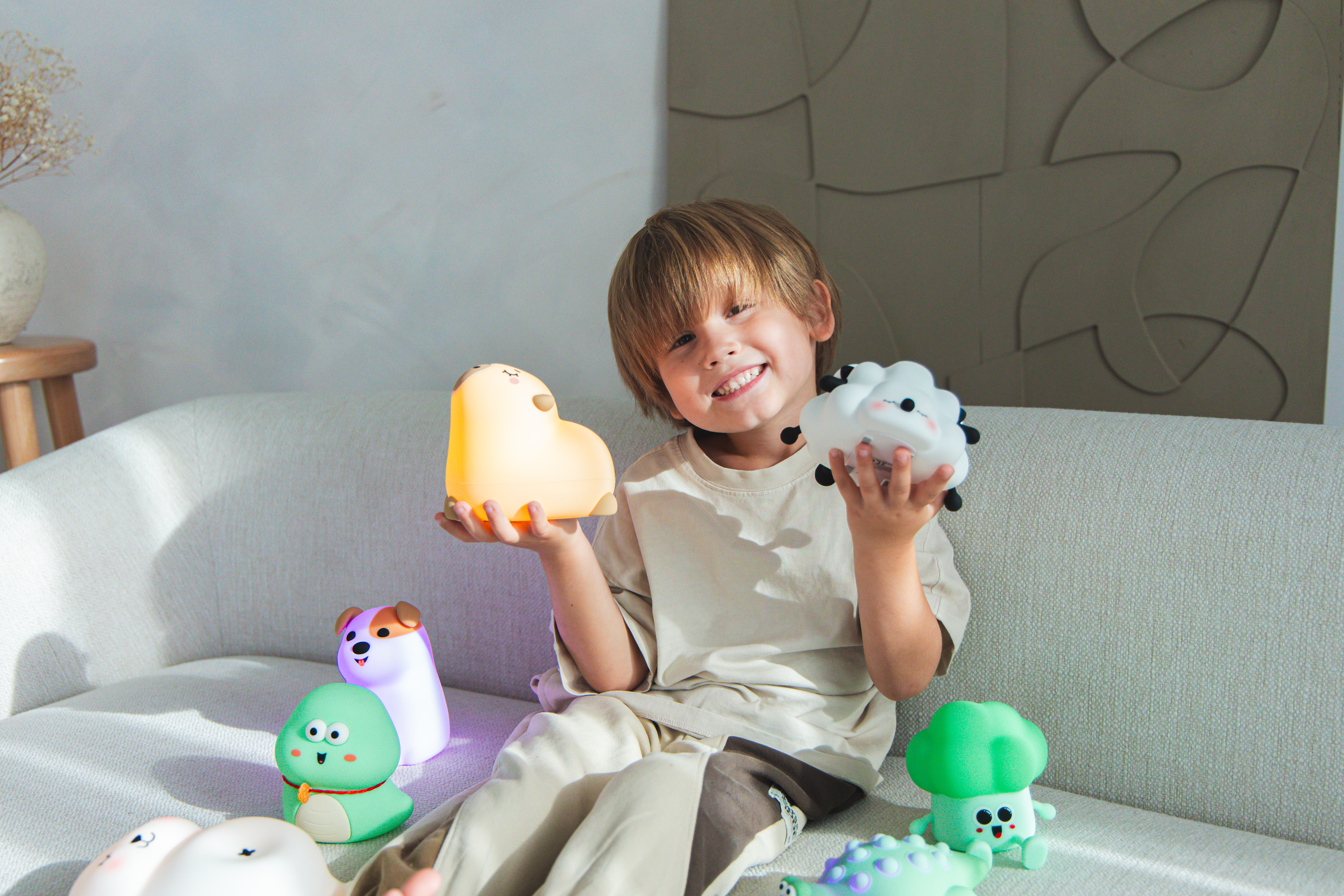 Uśmiechnięty chłopiec na kanapie trzyma żółtą i białą zabawkę. Na kanapie są inne zabawki. Tłem jest ściana i wazon.