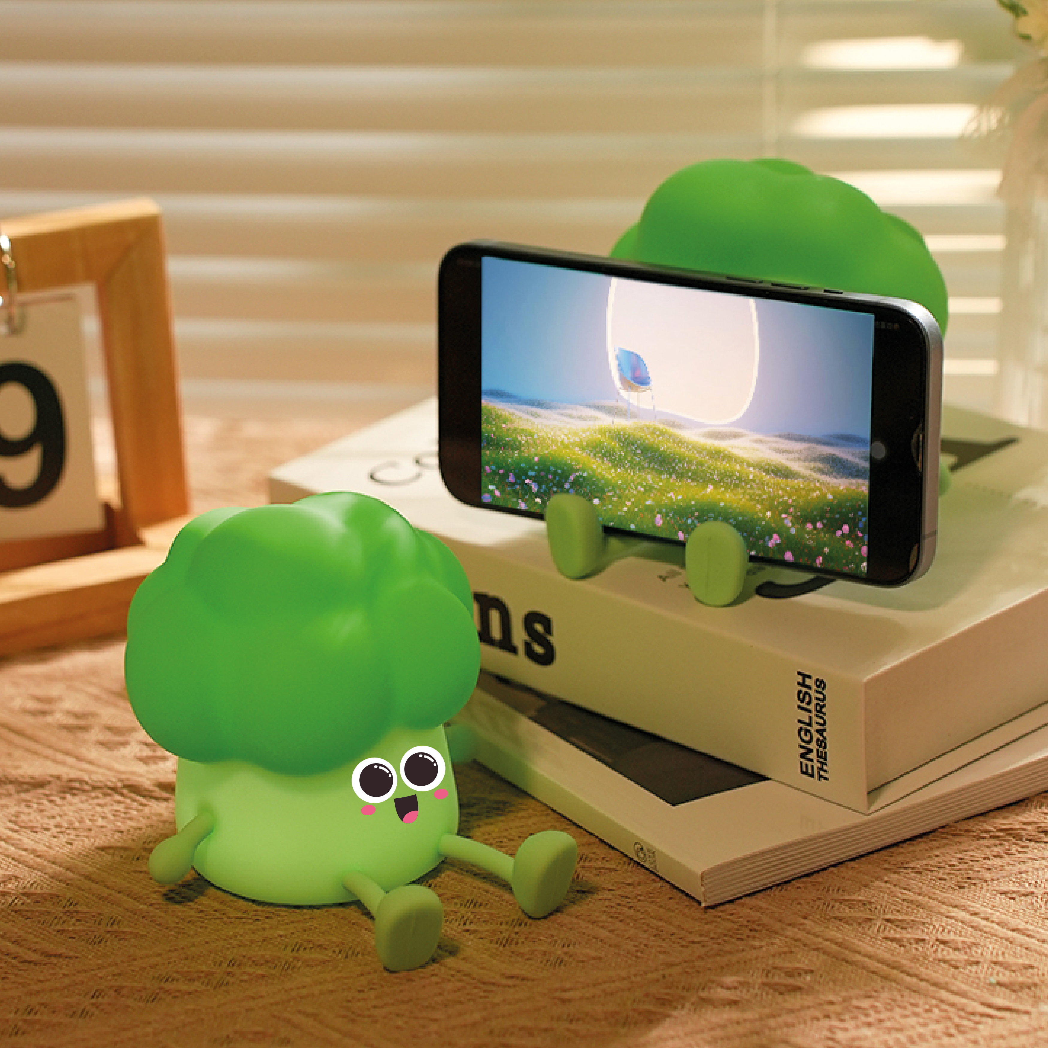 Kreskówkowy stojak na telefon w kształcie brokuła trzyma telefon, który wyświetla krajobraz. Drugi brokuł siedzi w pobliżu na książce.