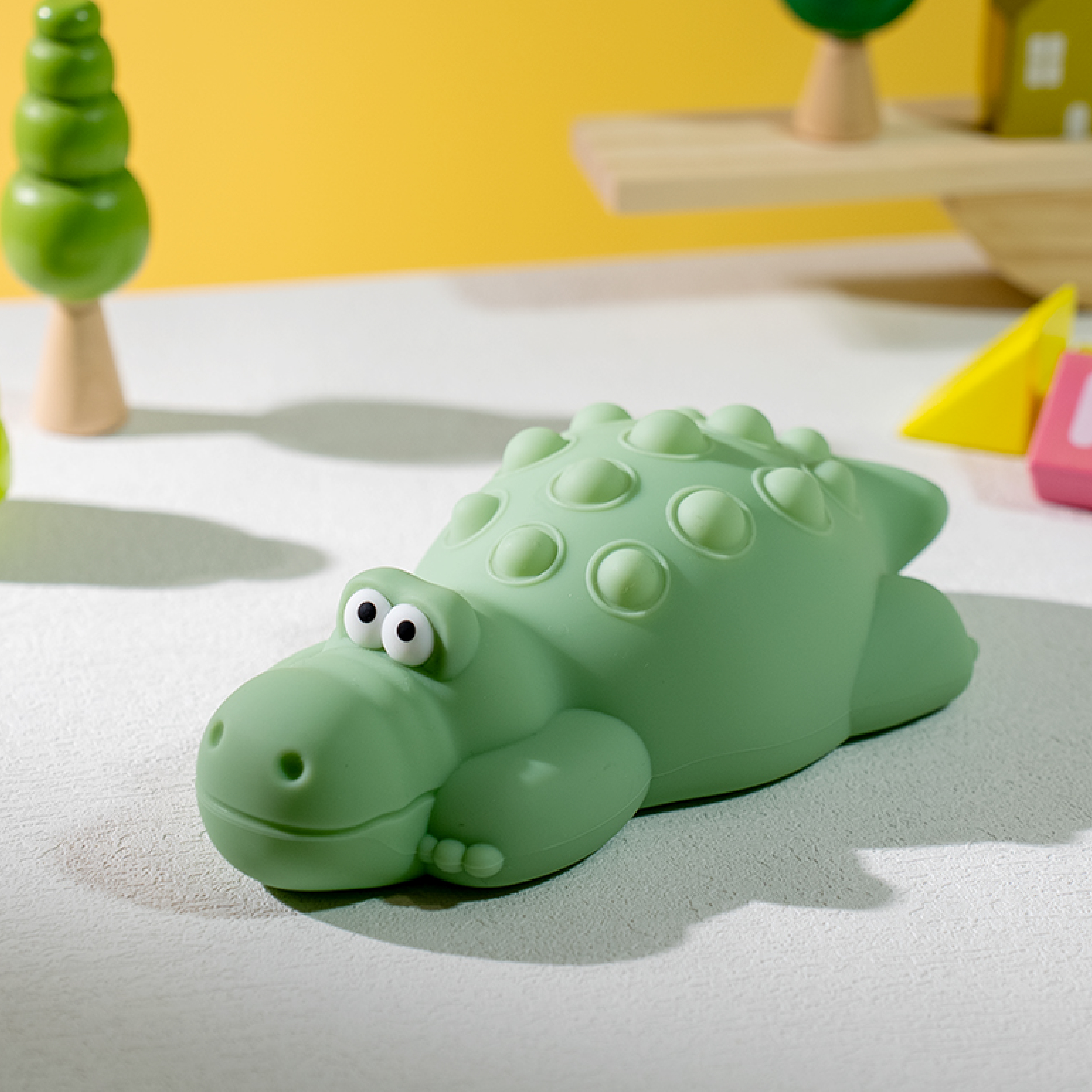 Zielona zabawka krokodyla na białej powierzchni z innymi zabawkami i żółtym tłem.