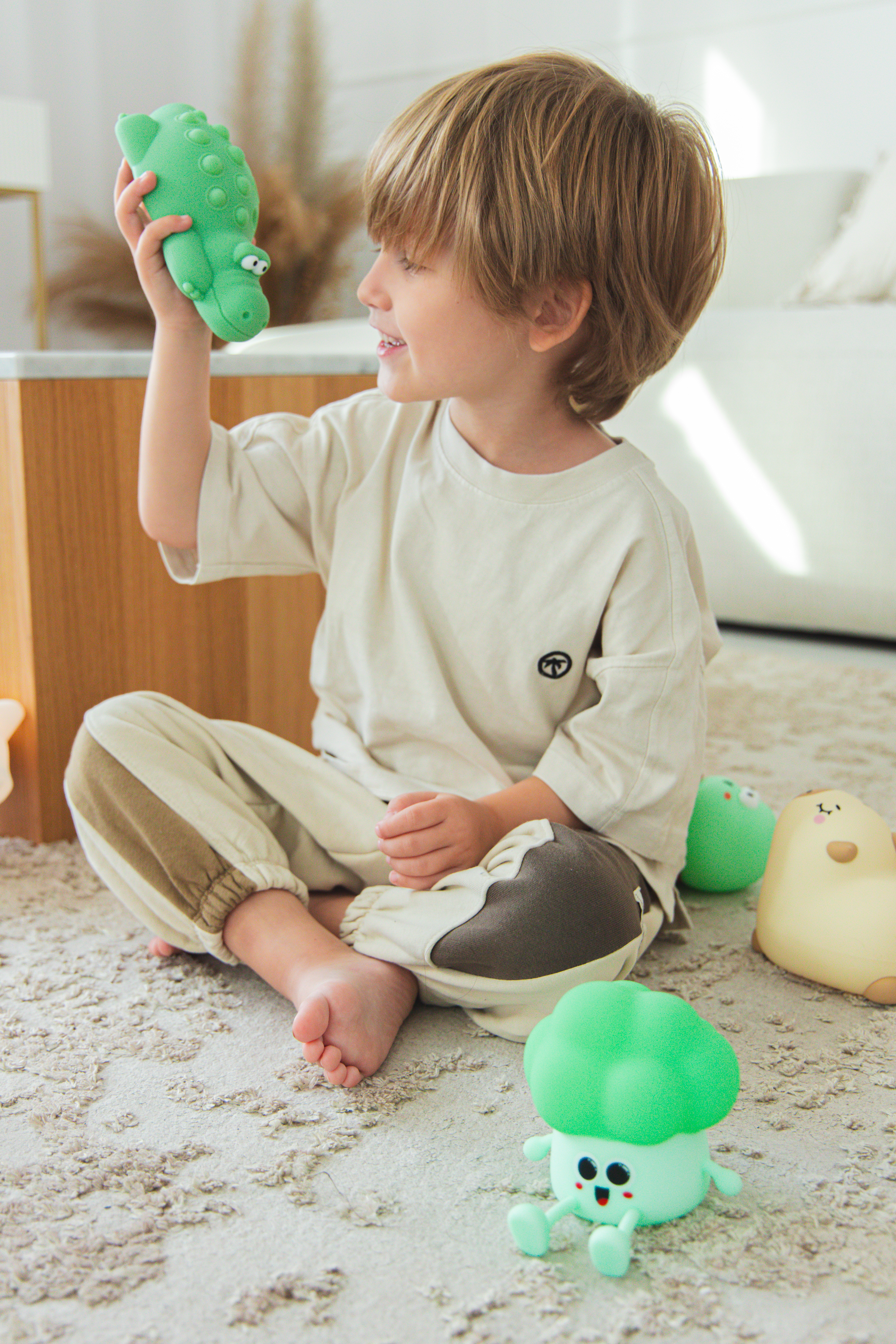Dziecko trzyma zielonego aligatora-zabawkę i się uśmiecha. Inne zabawki są na dywanie. Dziecko ma na sobie jasny strój.