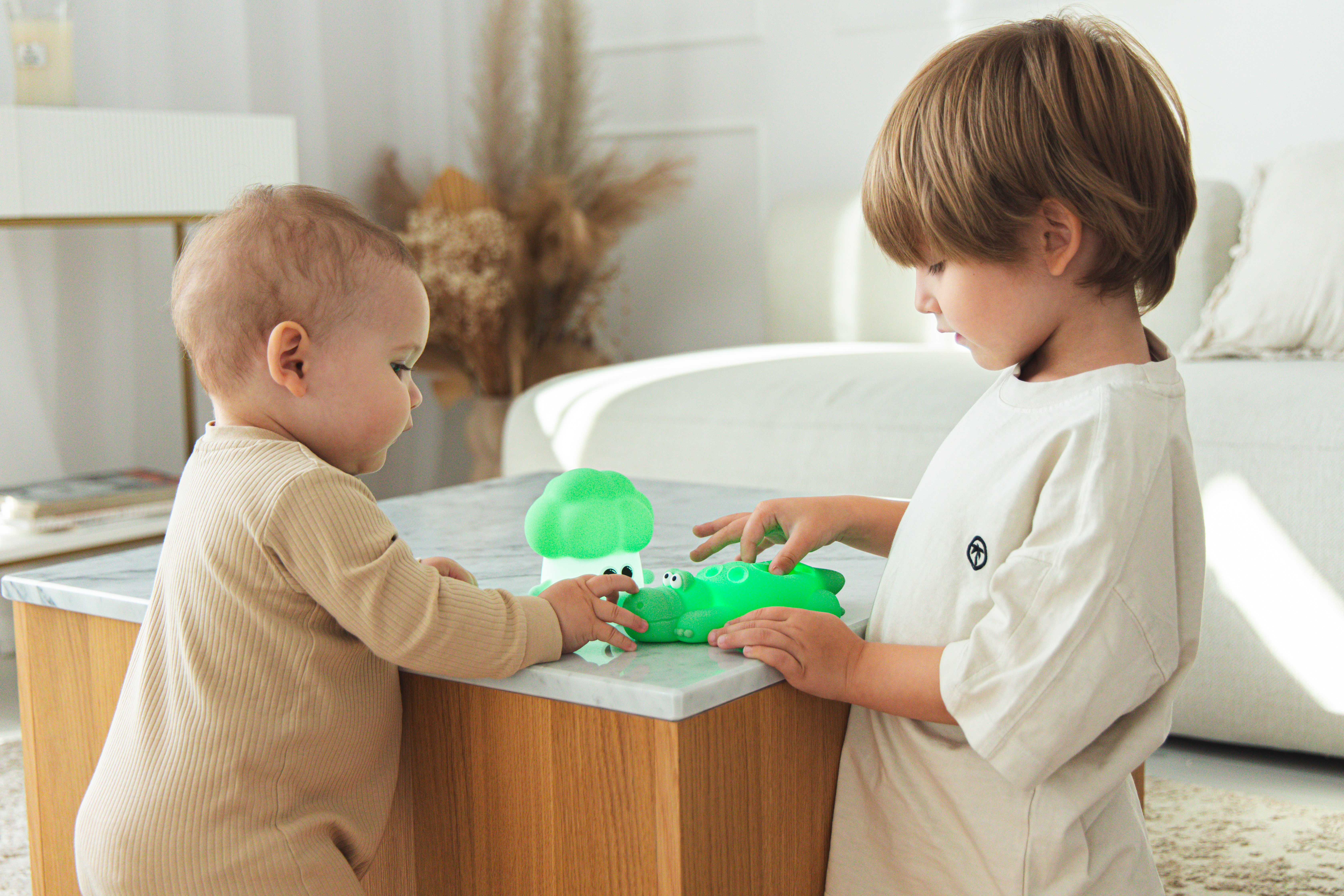 Dwoje dzieci bawi się zielonymi zabawkami na stole. Jedno to niemowlę, drugie chłopiec. Tłem jest salon.