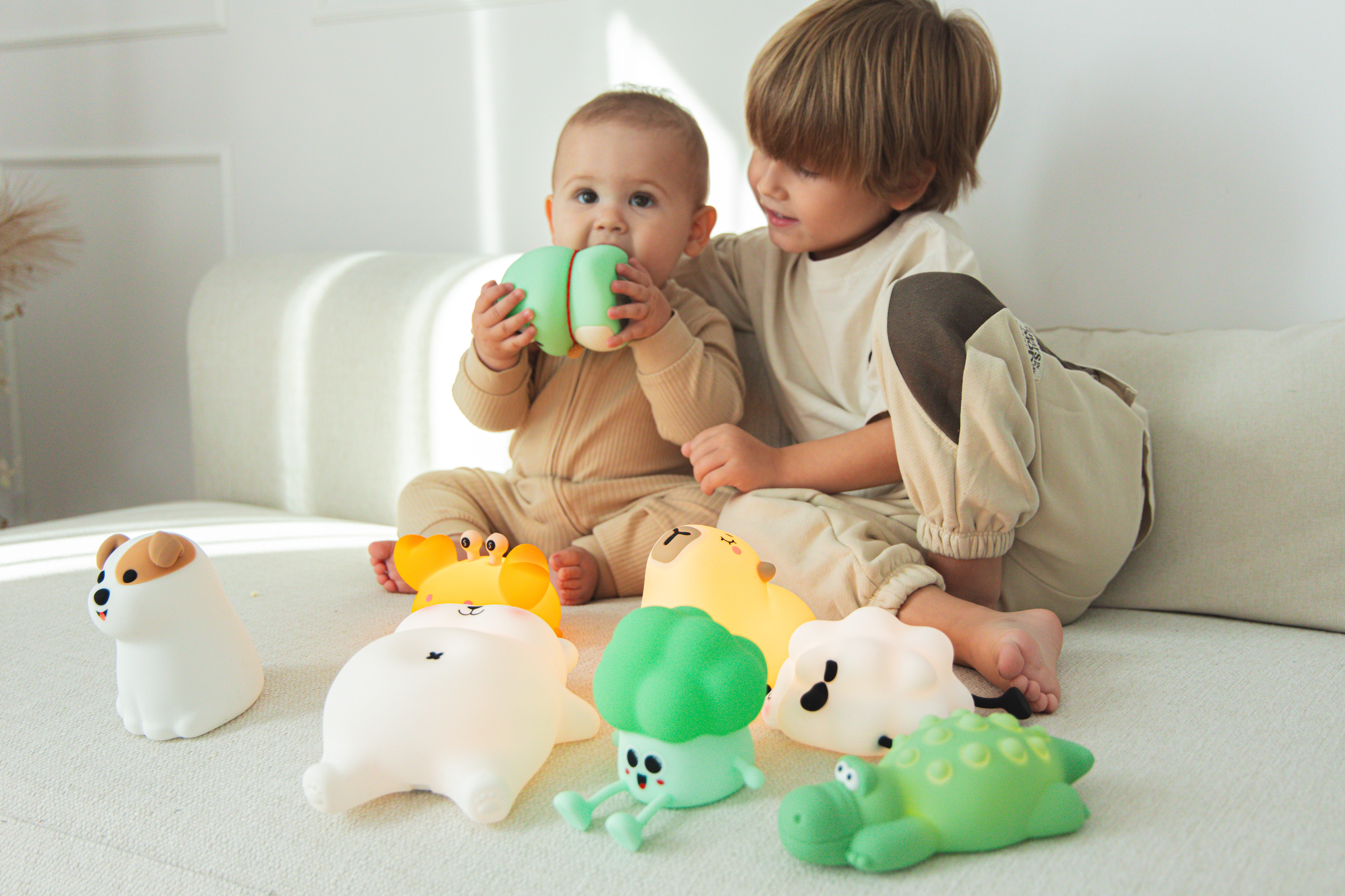 Niemowlę i chłopiec siedzą na kanapie z zabawkami. Niemowlę trzyma zieloną zabawkę. Kilka świecących zabawek znajduje się na pierwszym planie.