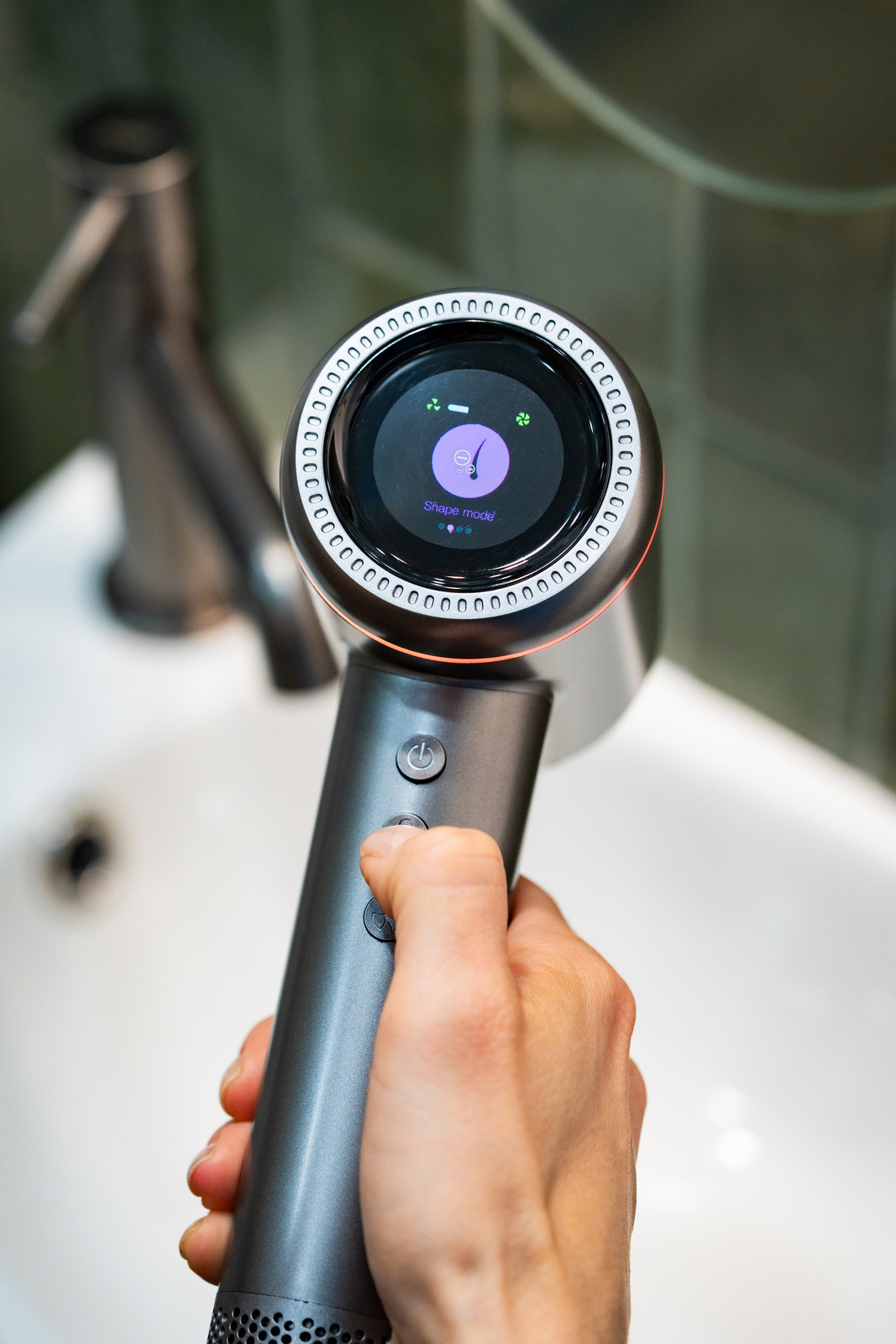 Dłoń trzyma szarą suszarkę do włosów z ekranem wyświetlającym 'Shape mode'. Umywalka i kran są na rozmytym tle.