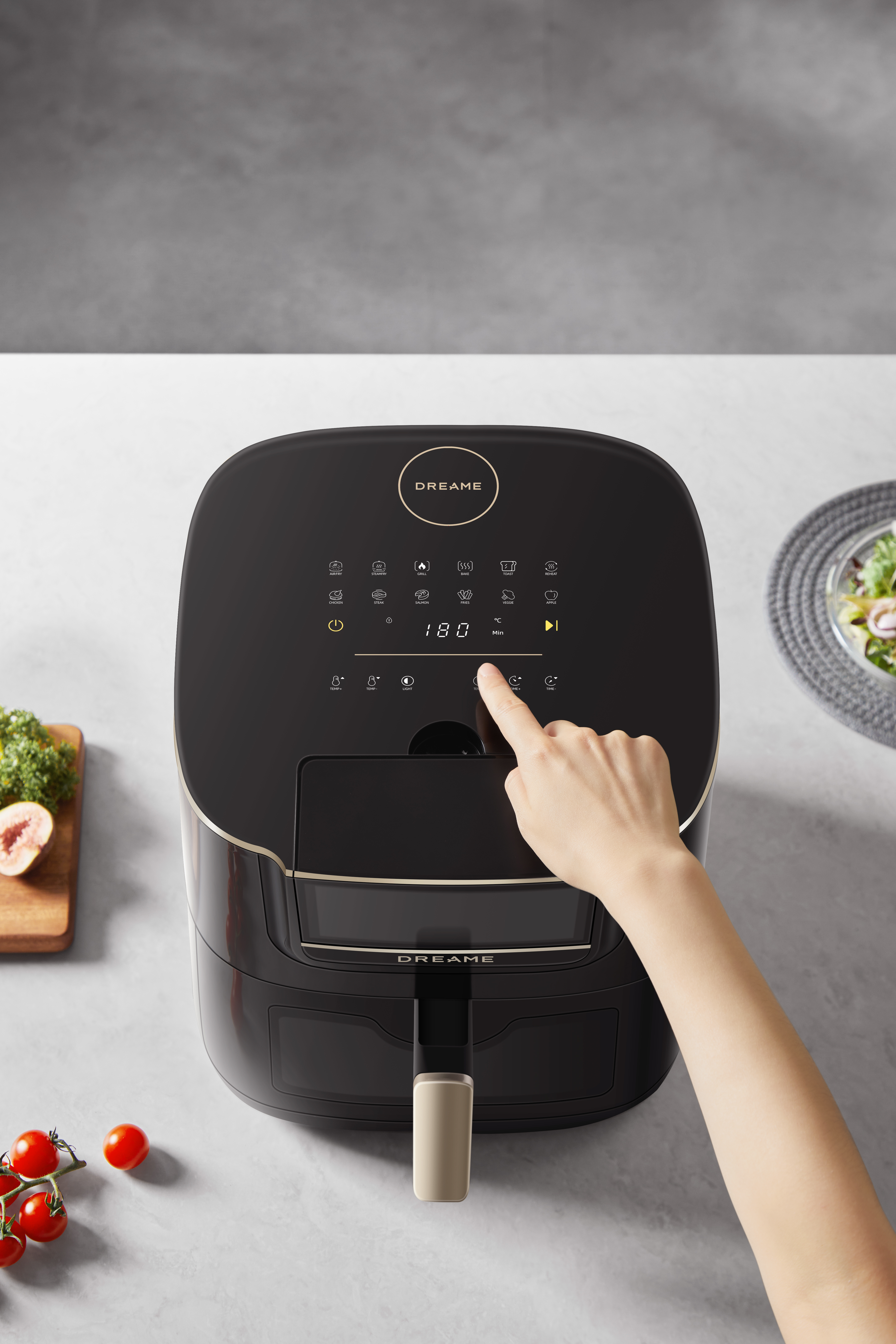 Ręka naciska przycisk na czarnej frytkownicy. Widoczne są również pomidory, figi i sałatka.