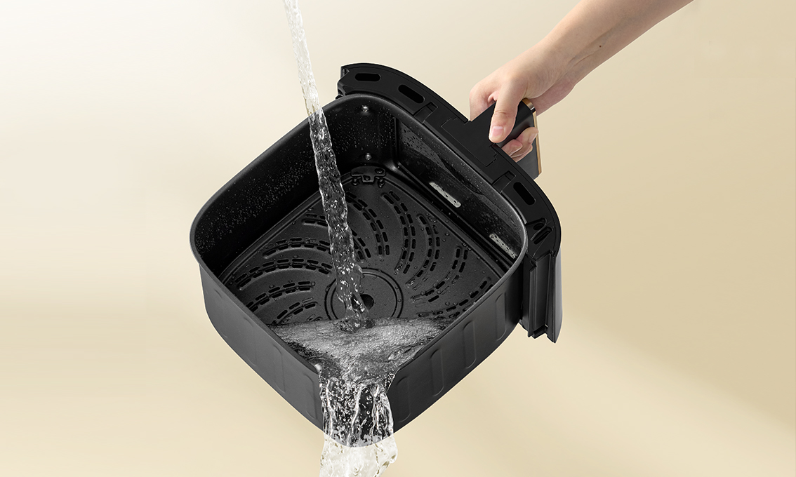 Kosz do frytkownicy powietrznej jest myty. Woda leje się z kranu do czarnego kosza trzymanego przez rękę. Beżowe tło.