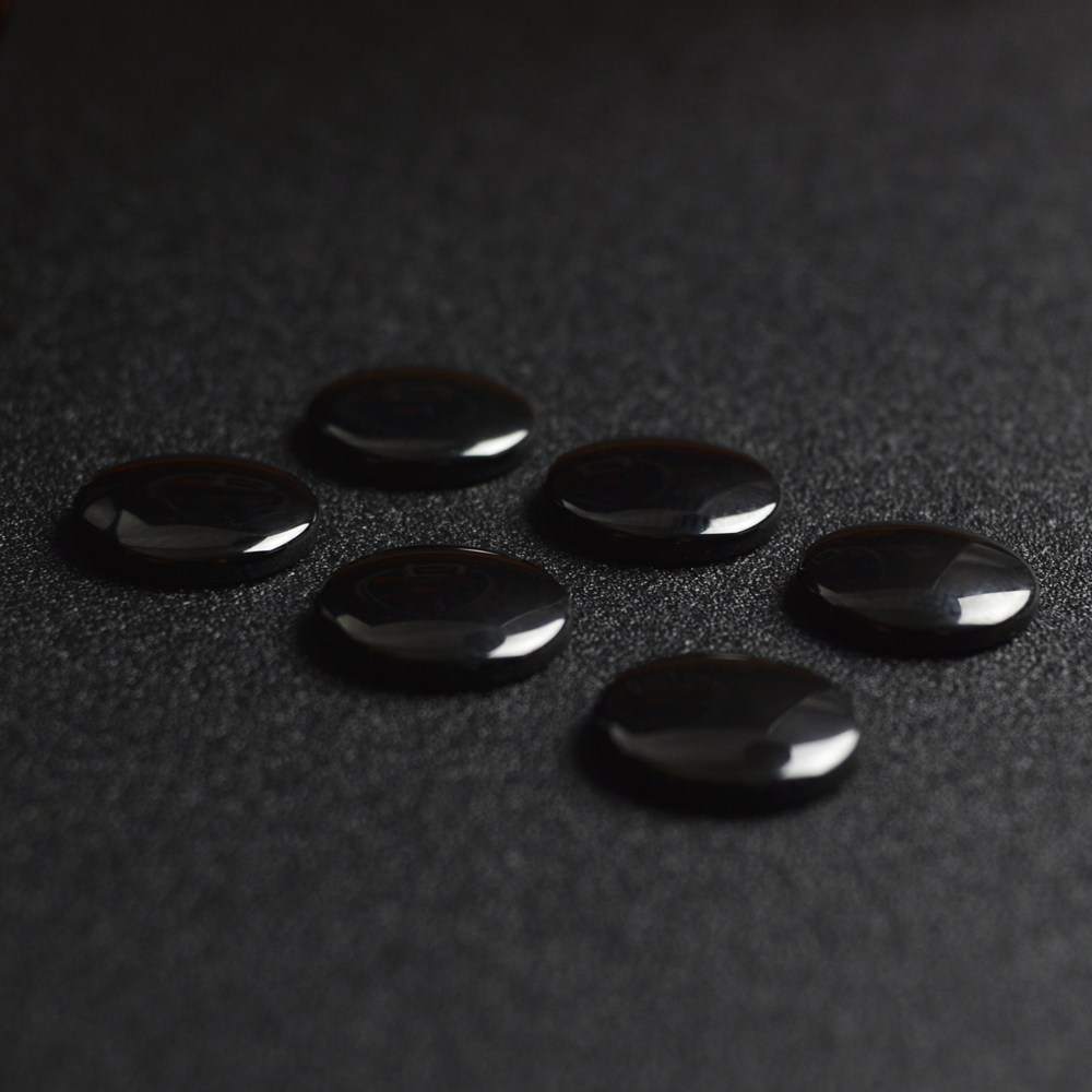 Sześć błyszczących, czarnych, owalnych kamieni ułożonych na teksturowanej, ciemnoszarej powierzchni. Na kamieniach widać odbicia.