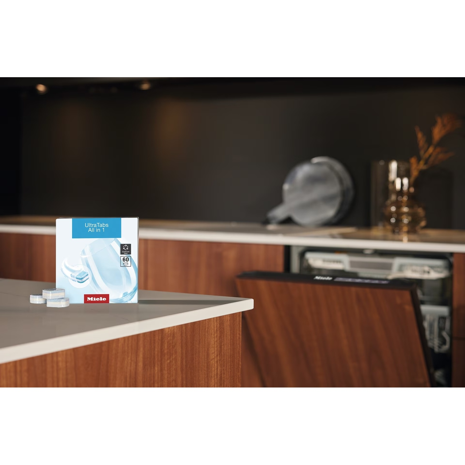 Una scatola di Miele UltraTabs All in 1 su un piano di lavoro, con tre pastiglie davanti. Una lavastoviglie sullo sfondo.
