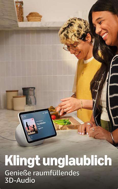 Zwei Frauen an einer Theke, lächelnd. Eine schneidet Gemüse. Ein Tablet zeigt eine Musik-App. Küchenhintergrund mit Regalen.