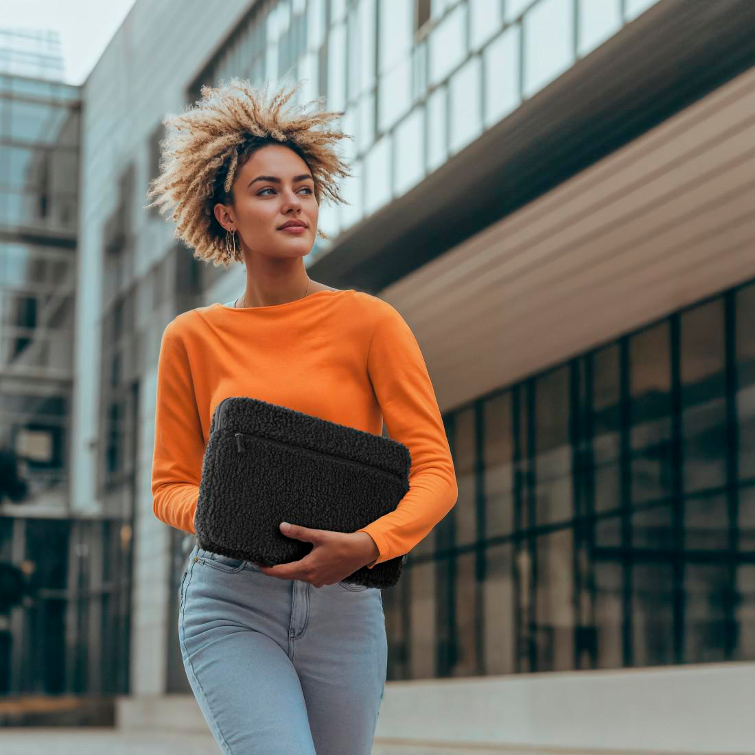 Frau in orangefarbenem Oberteil und Jeans, trägt eine schwarze, flauschige Laptoptasche, geht in der Nähe eines modernen Gebäudes.