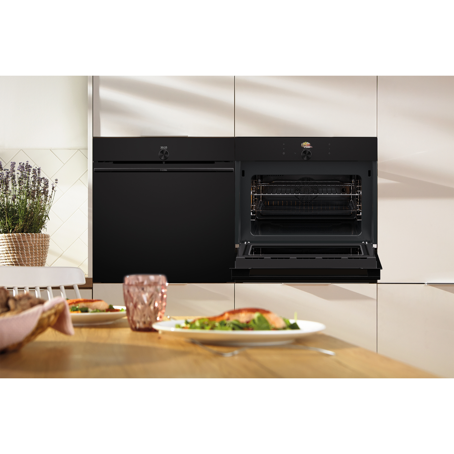 Een zwarte oven met de deur open, die de binnenkant laat zien. Een tafel is gedekt met borden eten. Een plant staat op de achtergrond.