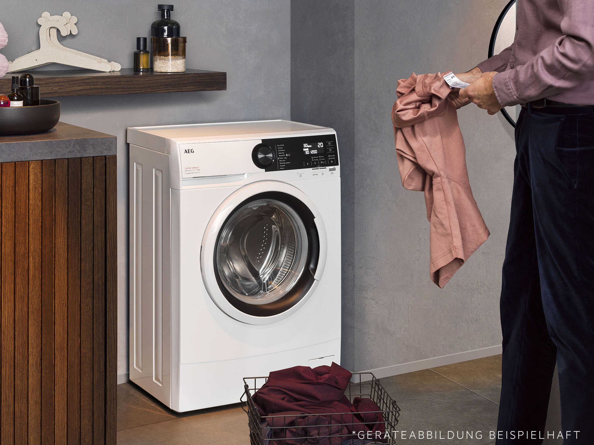 Eine weiße Waschmaschine steht neben einer Person, die ein rosa Hemd hält. Auf dem Boden steht ein Wäschekorb.