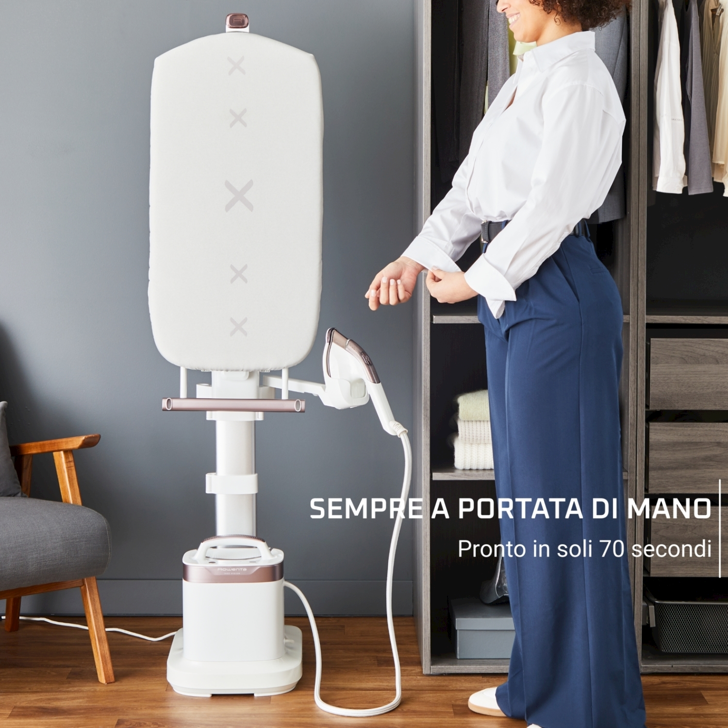 Donna che si rimbocca le maniche vicino a un vaporizzatore. Il vaporizzatore è bianco con un asse da stiro grigio. I vestiti sono sullo sfondo.