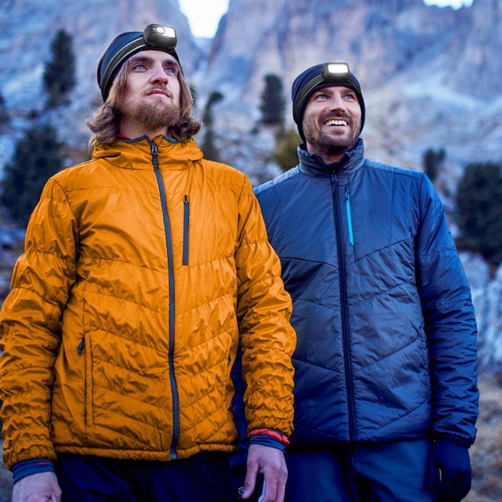 Dwóch mężczyzn w czołówkach i kurtkach spogląda na góry. Jedna kurtka jest pomarańczowa, druga niebieska. Tło przedstawia góry.