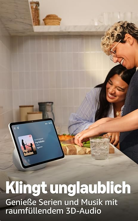 Zwei Personen lächeln auf ein Tablet, das in einer Küche Musik abspielt. Essen und ein Glas Wasser sind auf der Theke zu sehen.