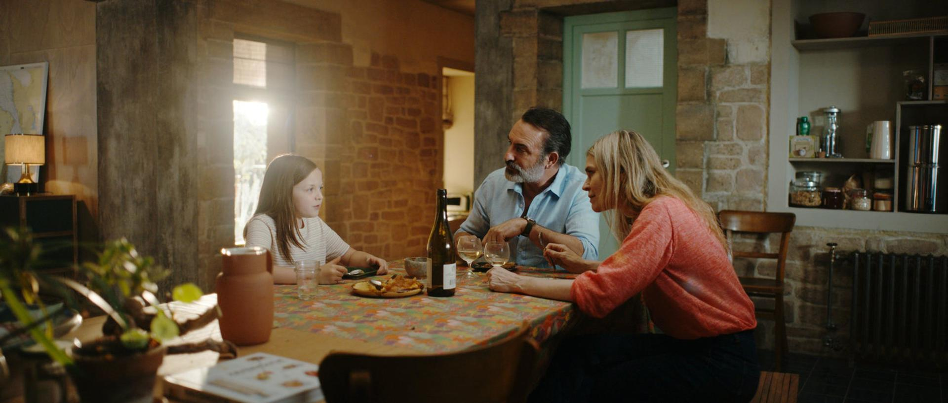 Familie am Tisch, ein Mädchen und zwei Erwachsene, essen und reden. Eine Weinflasche und Gläser stehen auf dem Tisch.