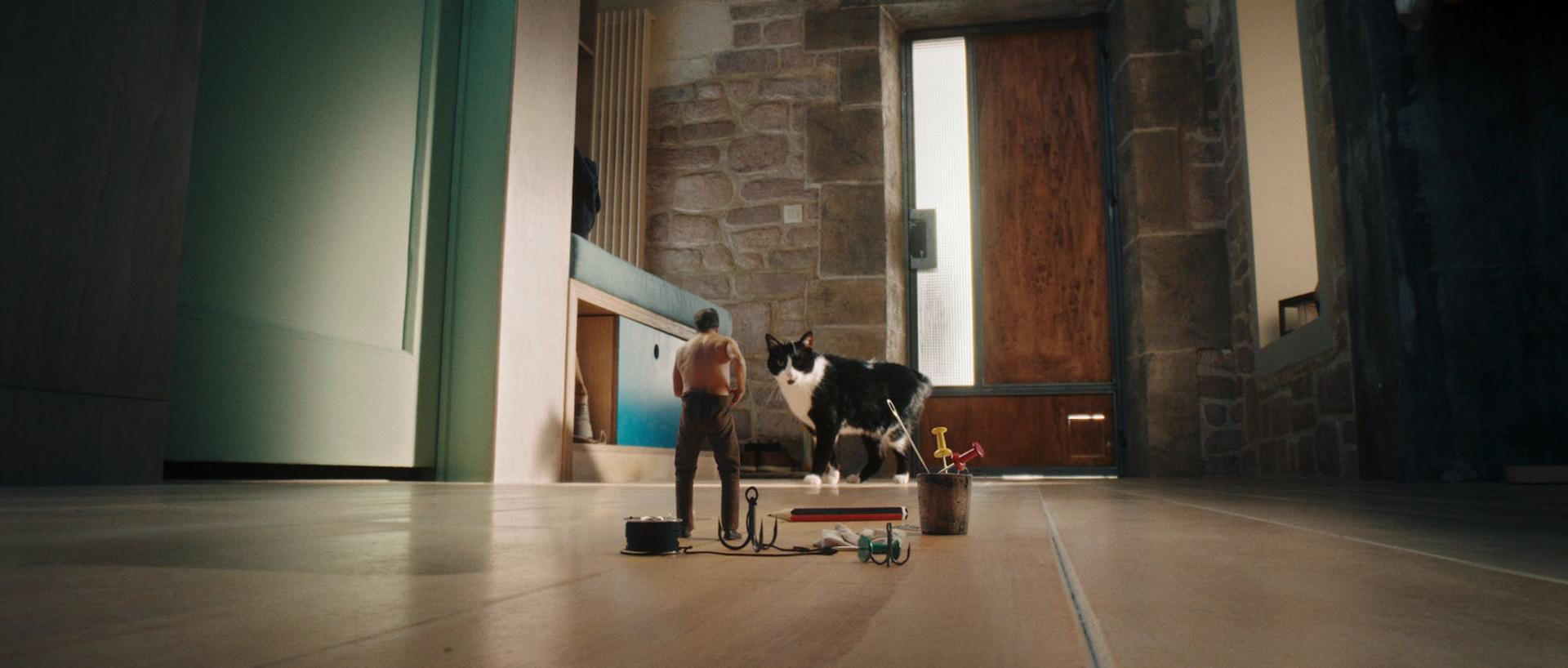 Ein Mann steht mit Angelausrüstung, einer schwarz-weißen Katze und einer Holztür. Die Szene spielt sich in Innenräumen mit Steinmauern ab.