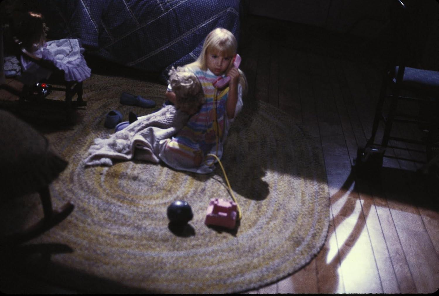 Ein Mädchen mit einer Puppe sitzt auf einem Teppich und telefoniert mit einem rosa Telefon. Eine Puppe in einem Schaukelstuhl und ein schwarzer Ball sind in der Nähe.
