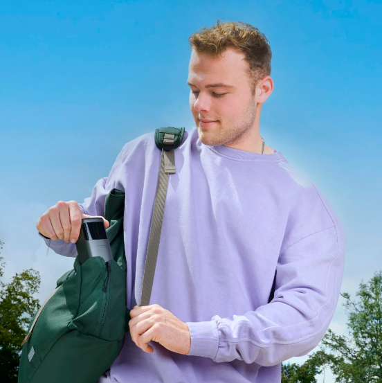 Młody mężczyzna w fioletowym swetrze zagląda do zielonego plecaka. Jest na zewnątrz pod błękitnym niebem, z drzewami w tle.