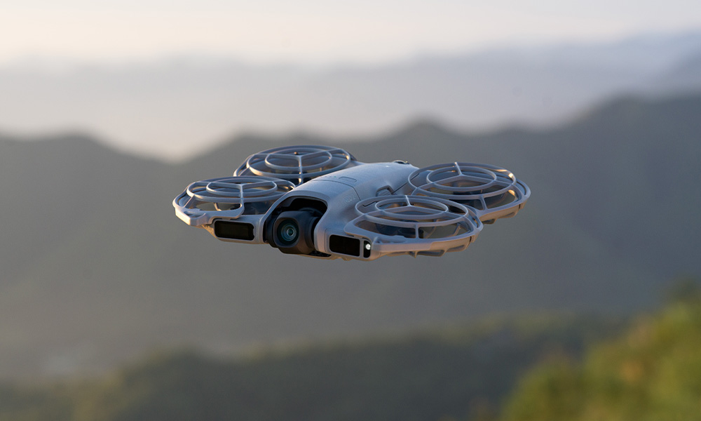 Dron leci w powietrzu. Ma cztery śmigła i kamerę. Tło przedstawia rozmyte góry i niebo.