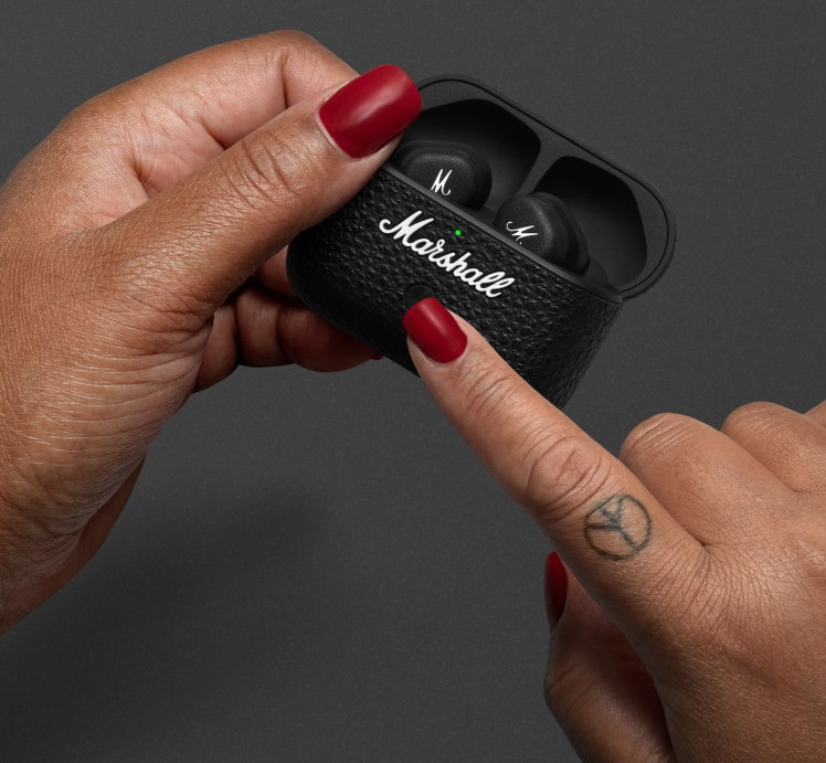 Dłonie trzymające czarne etui na słuchawki Marshall ze słuchawkami w środku. Czerwony lakier do paznokci. Tatuaż ze znakiem pokoju na palcu. Zielone światło na etui.