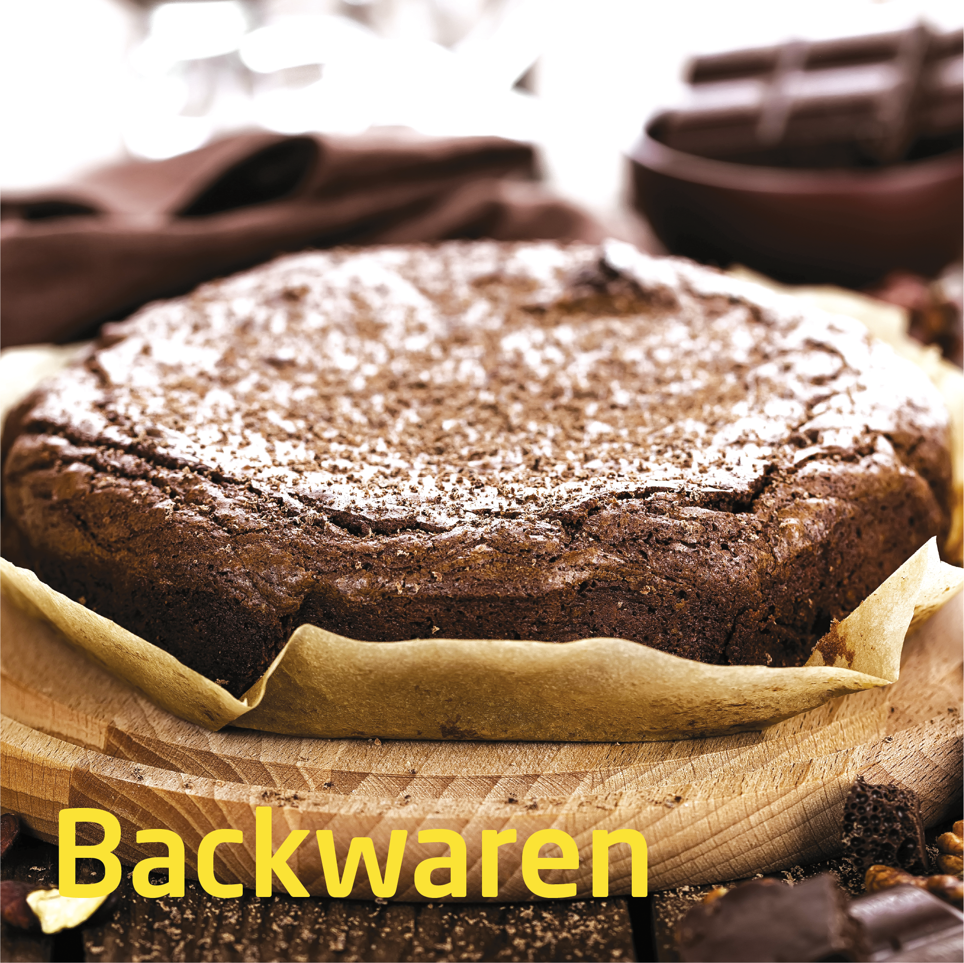 Ein runder Schokoladenkuchen mit Puderzucker bestäubt, auf einem Holzbrett. Zutaten wie Schokolade und Nüsse sind dabei.