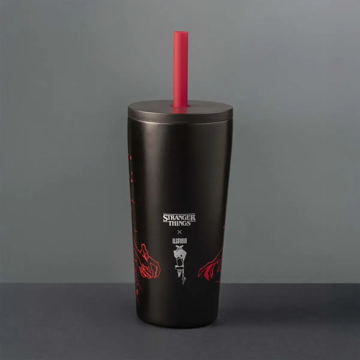 Fekete pohár piros szívószállal, 'Stranger Things' logóval és piros grafikus elemekkel. Szürke háttér.