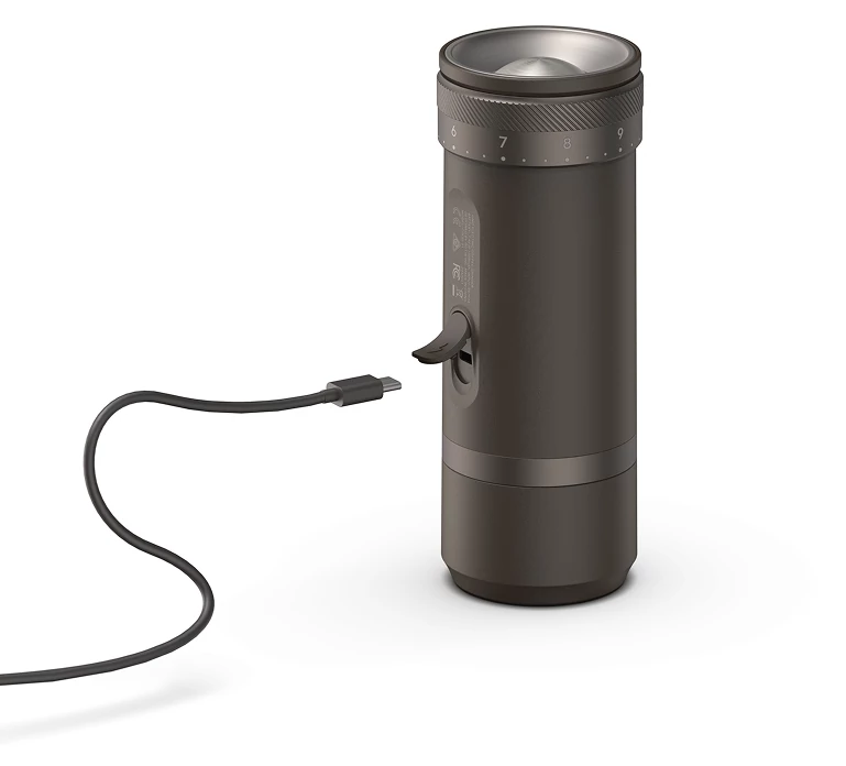 Egy hengeres kávéőrlő, csatlakoztatott USB-C kábellel. Sötétszürke, 6-9 számokkal a tetején. USB port az oldalon.
