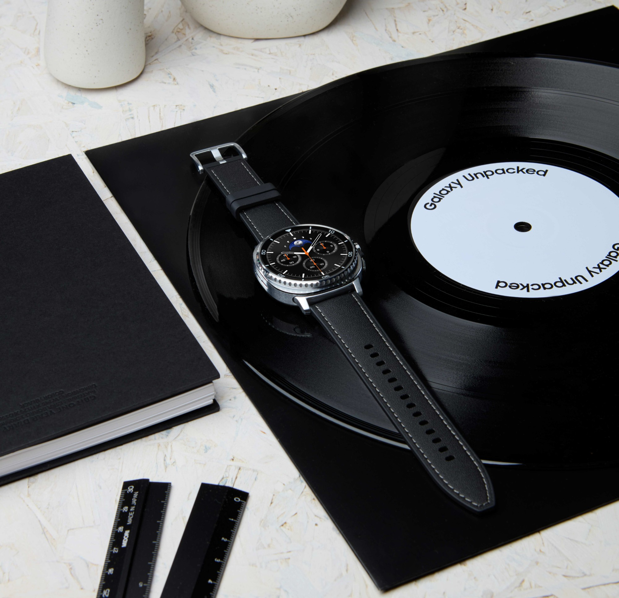 Zegarek z czarnym paskiem leży na płycie winylowej, obok czarnego notatnika i linijek. Tło jest białe.