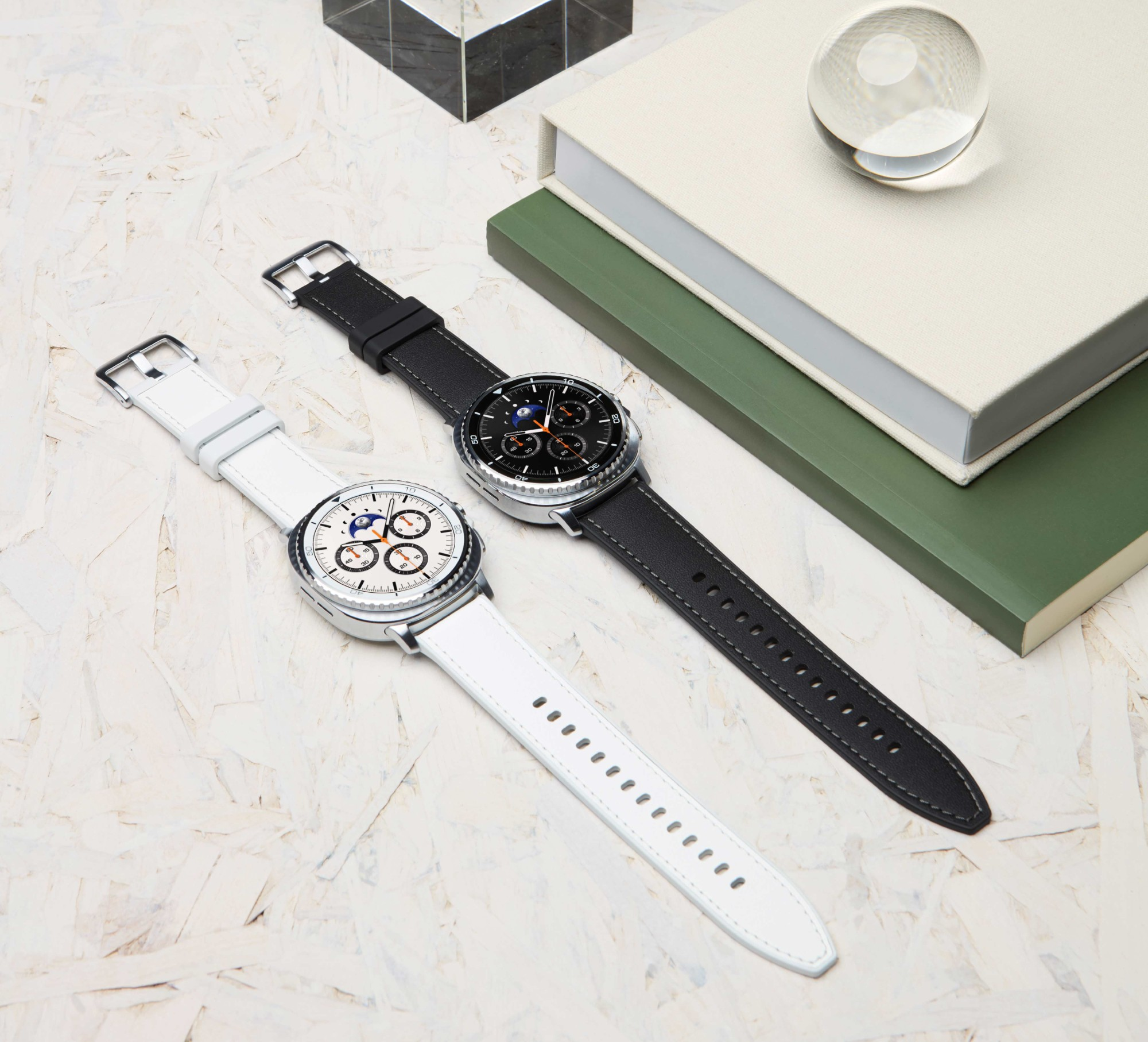 Dwa smartwatche, jeden biały pasek, jeden czarny pasek. Szklana kula i książki w tle.