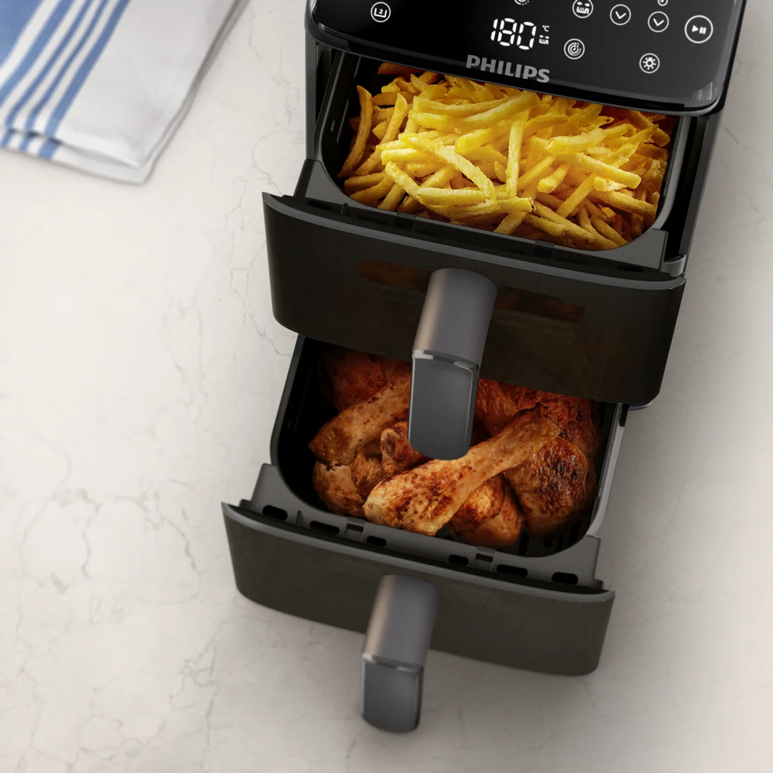 Friteuse à air Philips noire avec frites dans le bac supérieur, poulet cuit dans le bac inférieur, sur une surface en marbre blanc.
