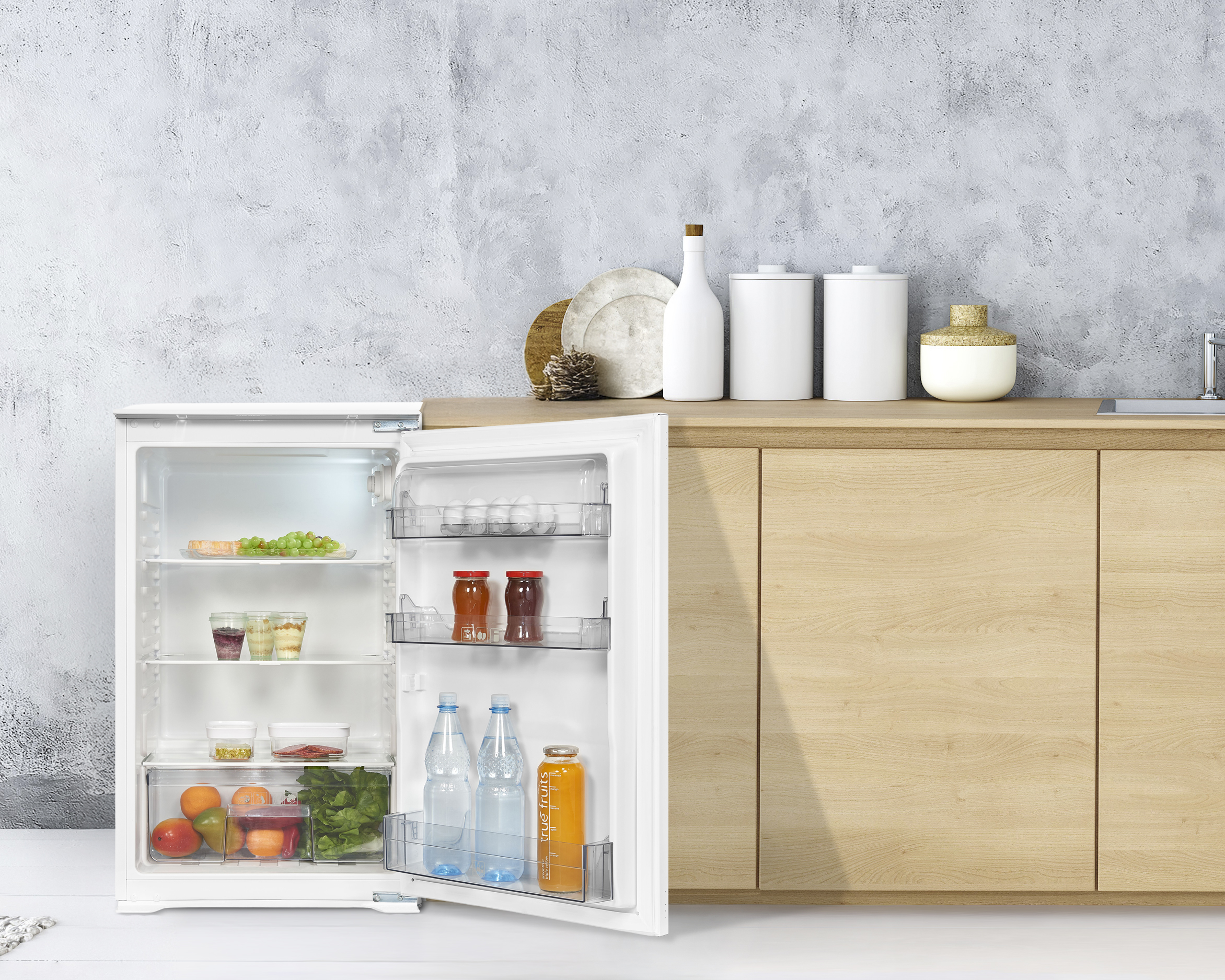 Offener Kühlschrank mit Lebensmitteln und Getränken. Hintergrund: Theke mit Gläsern und Holzschränken.