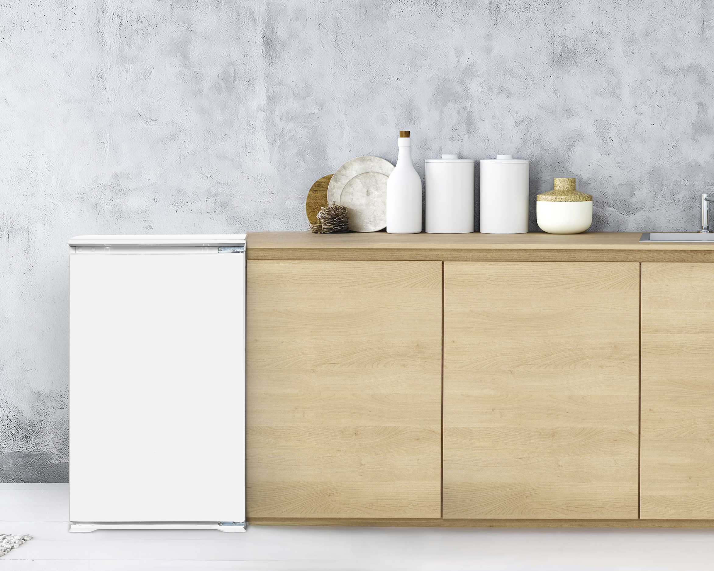 Ein weißer Kühlschrank steht neben einem Holzschrank mit weißen Gläsern und einer Flasche darauf, vor einer grauen Wand.
