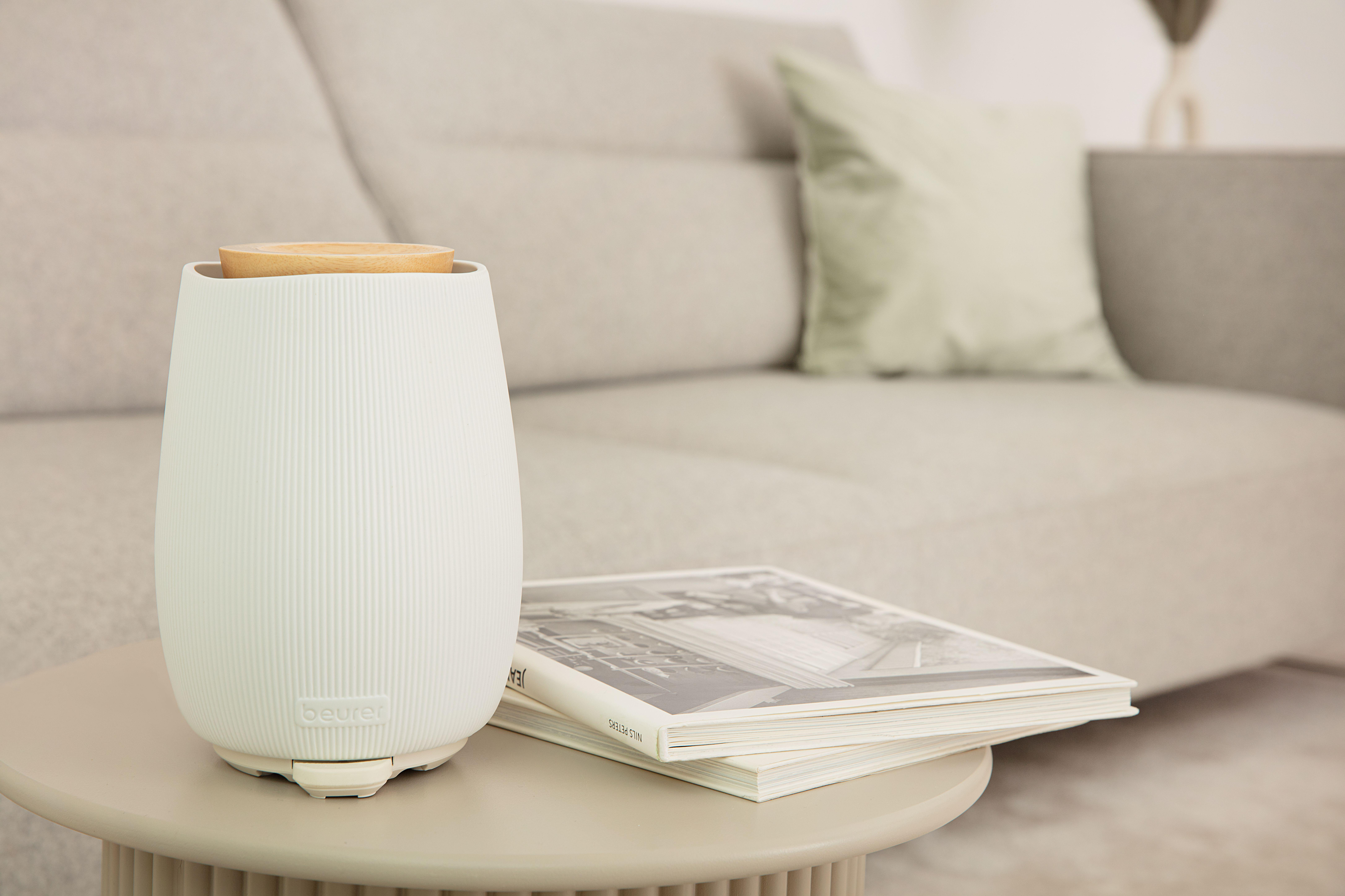 Ein weißer Diffusor mit Holzaufsatz steht auf einem Tisch. Im Hintergrund sind ein Magazin und ein Sofa mit Kissen.