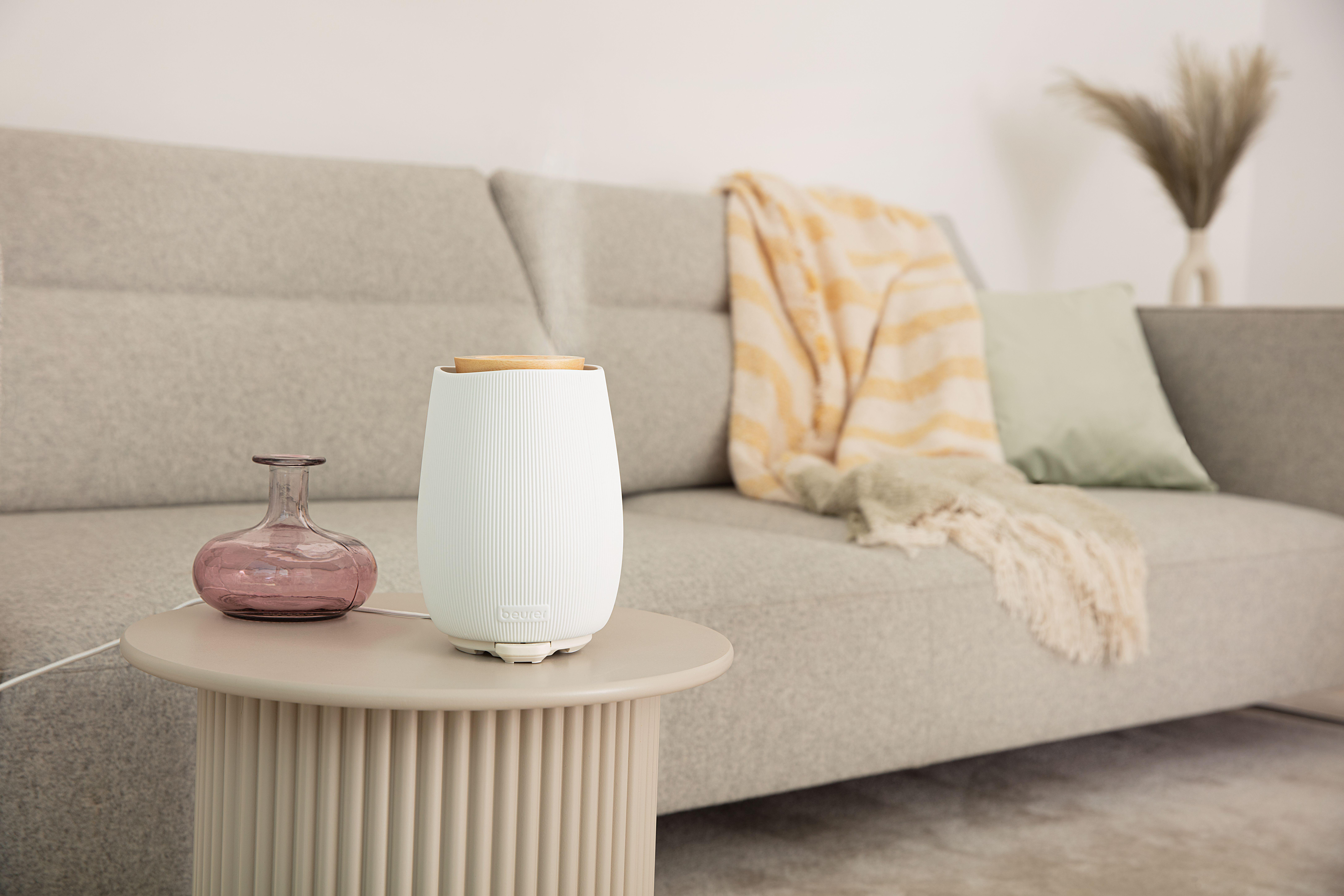 Ein weißer Diffusor, eine rosa Vase, ein beiger Tisch und ein graues Sofa mit gelber Decke und grünem Kissen.