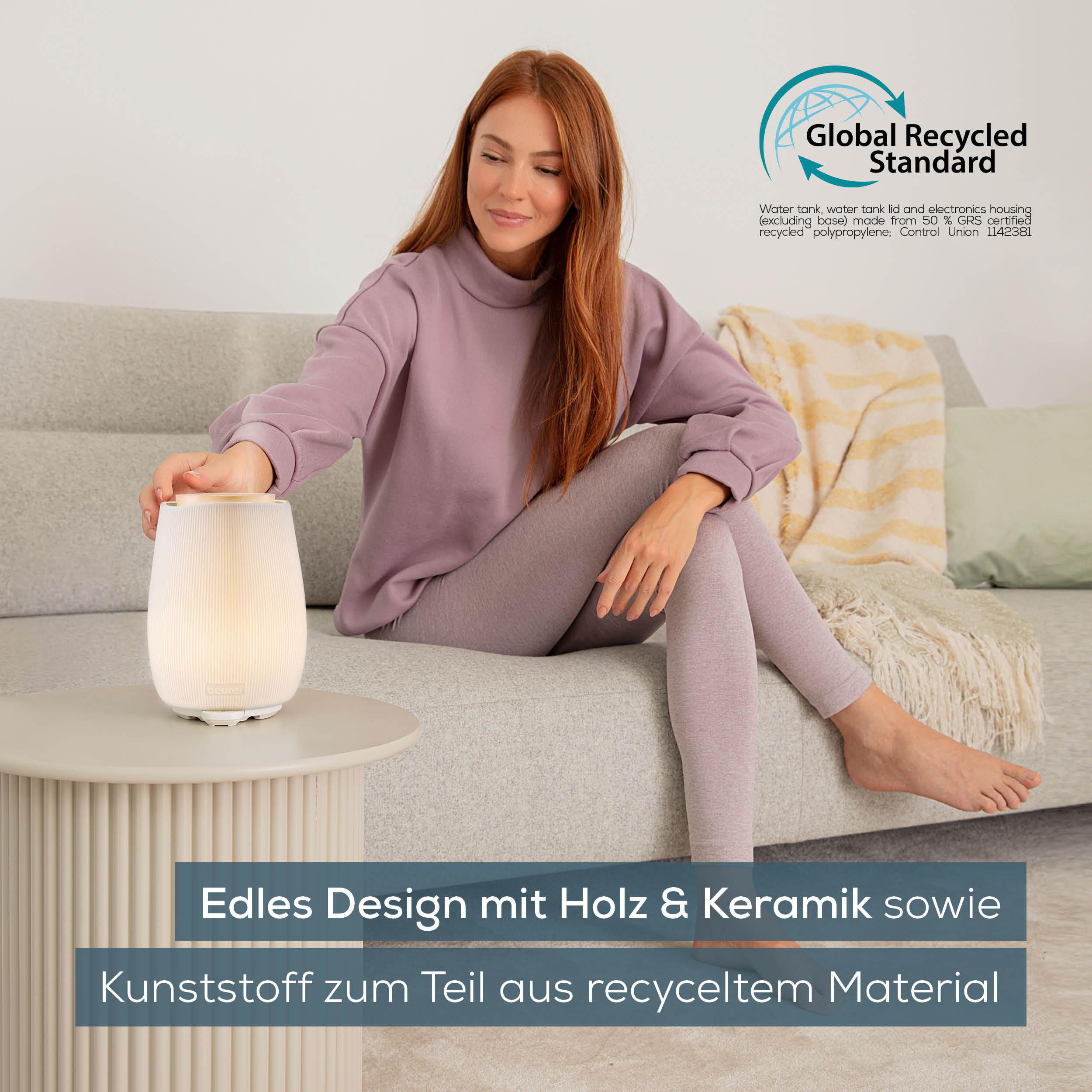 Frau in lila Outfit sitzt auf einem Sofa und berührt einen leuchtenden Aromadiffusor. Text: Edles Design mit Holz & Keramik sowie Kunststoff.