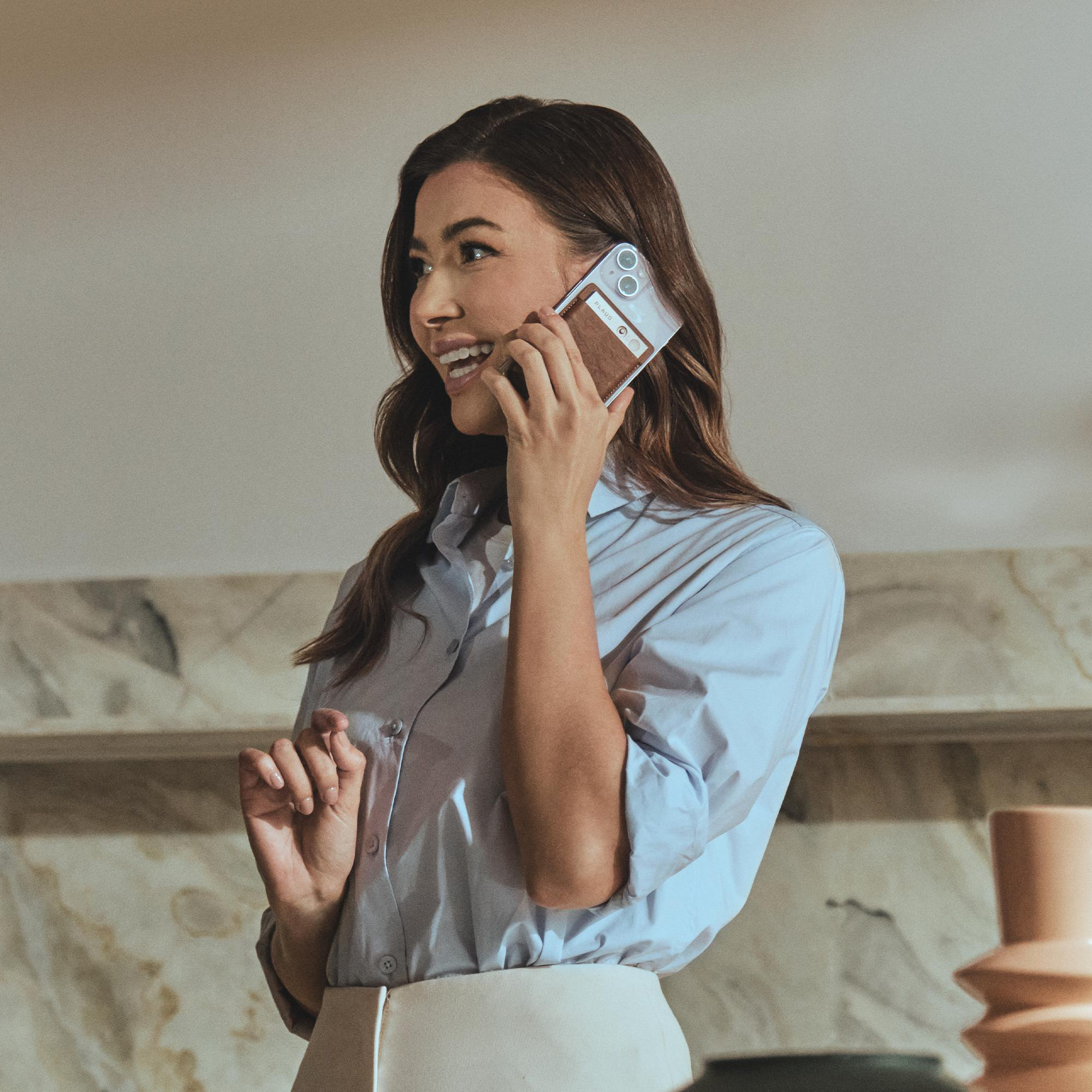 Eine Frau telefoniert lächelnd. Sie hält ein Telefon mit braunem Portemonnaie-Etui. Sie trägt ein hellblaues Hemd und einen weißen Rock.